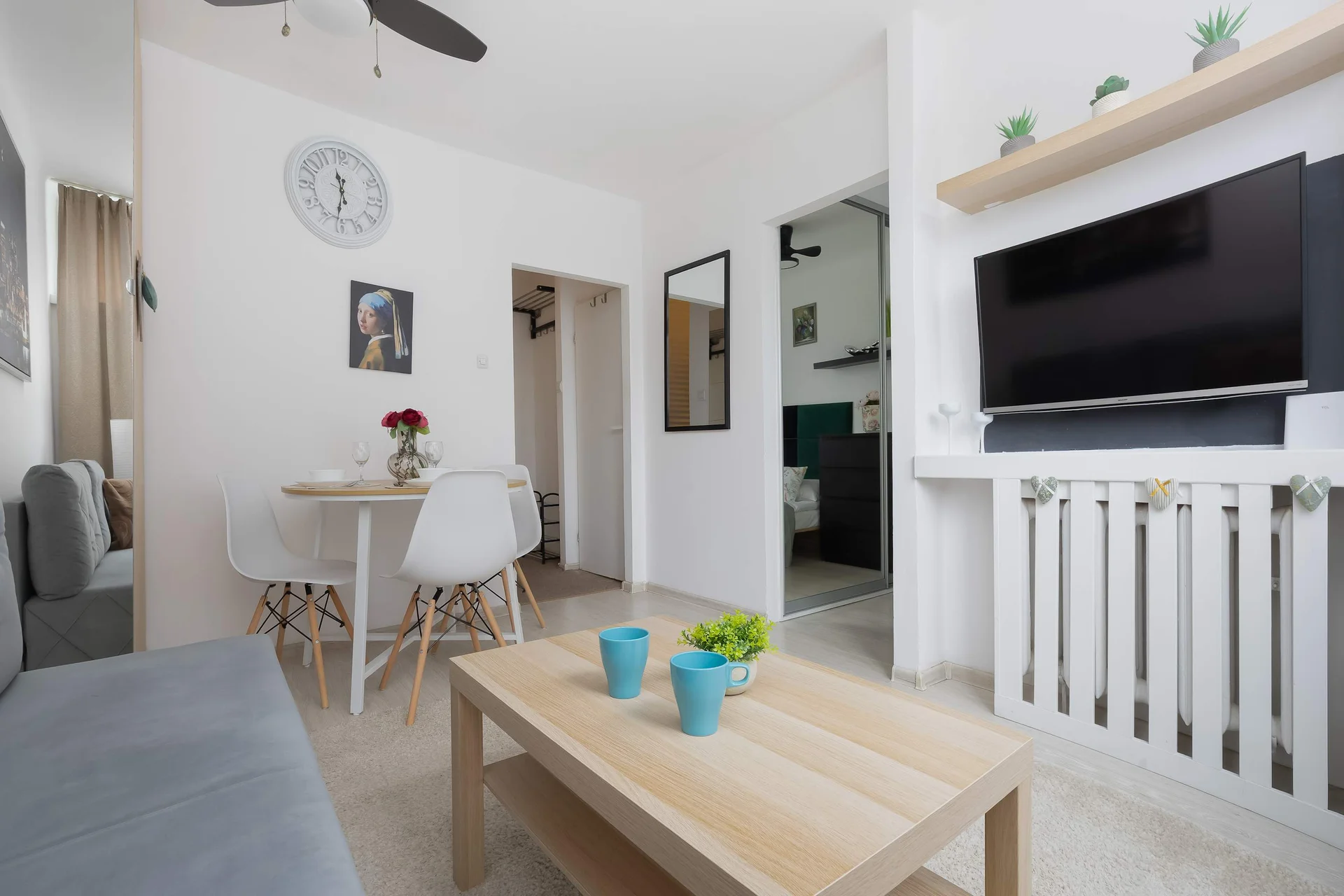 A cozy living room view features a wooden coffee table in the foreground adorned with two blue cups. In the background, a small white dining table with chairs sits near a wall-mounted flat-screen television.