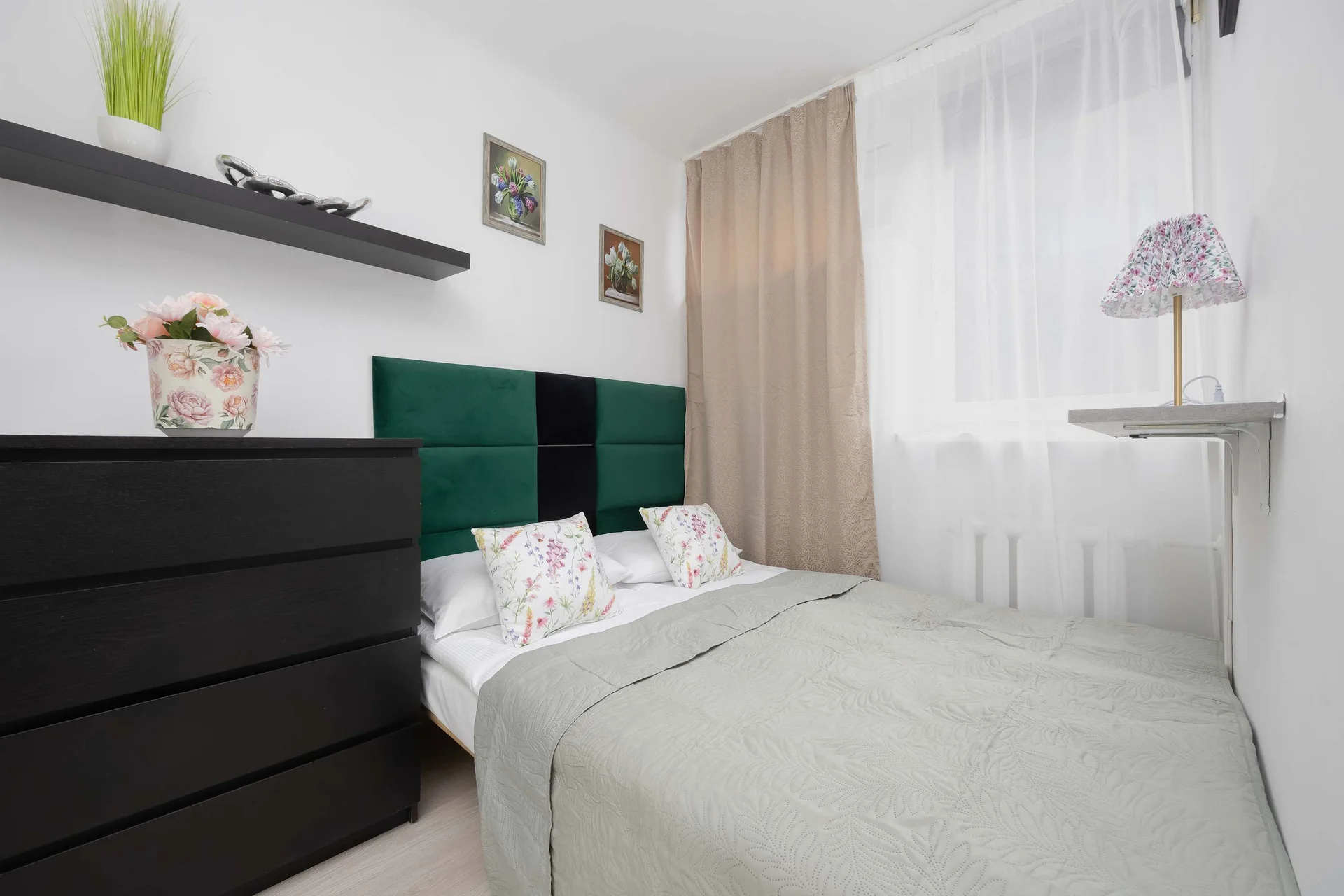 A neatly arranged bedroom showcases a double bed made up with a soft gray coverlet and accent pillows. Beside the bed stands a tall black chest of drawers against a clean white wall.