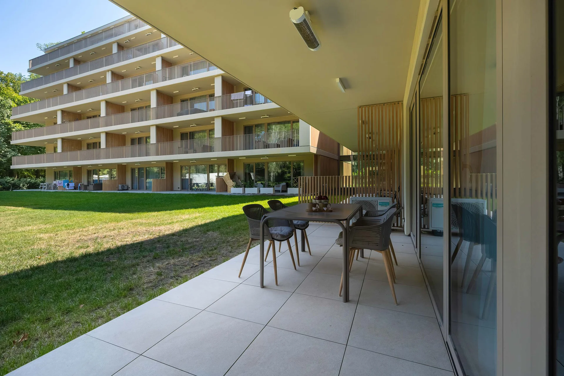 A spacious covered terrace offers a dining table with chairs overlooking a landscaped courtyard and modern apartment buildings. The outdoor space is ideal for relaxing or enjoying meals.