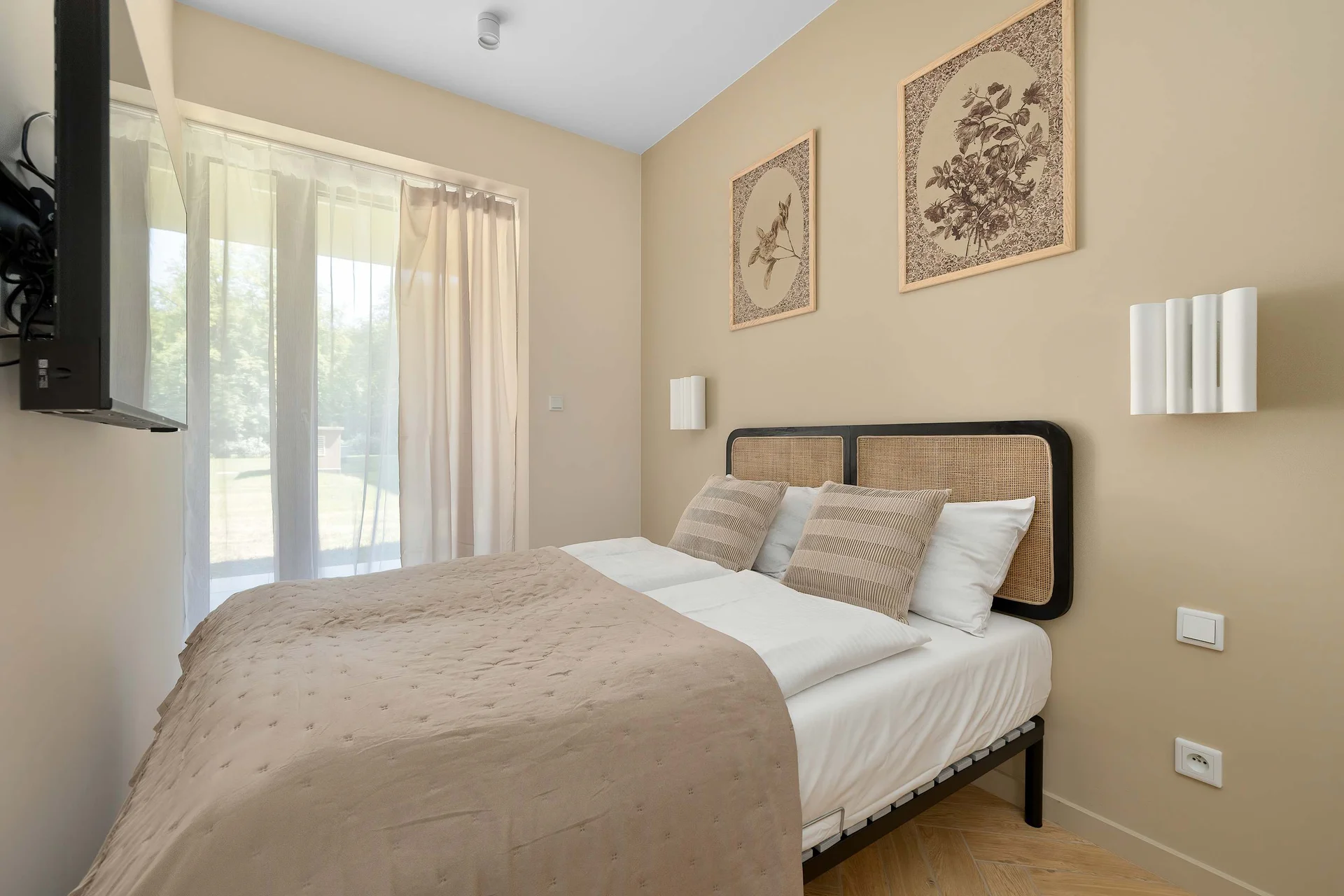 A bright bedroom features a double bed with beige bedding, matching bedside tables, and soft natural light streaming through large windows. Neutral tones create a warm and relaxing atmosphere.