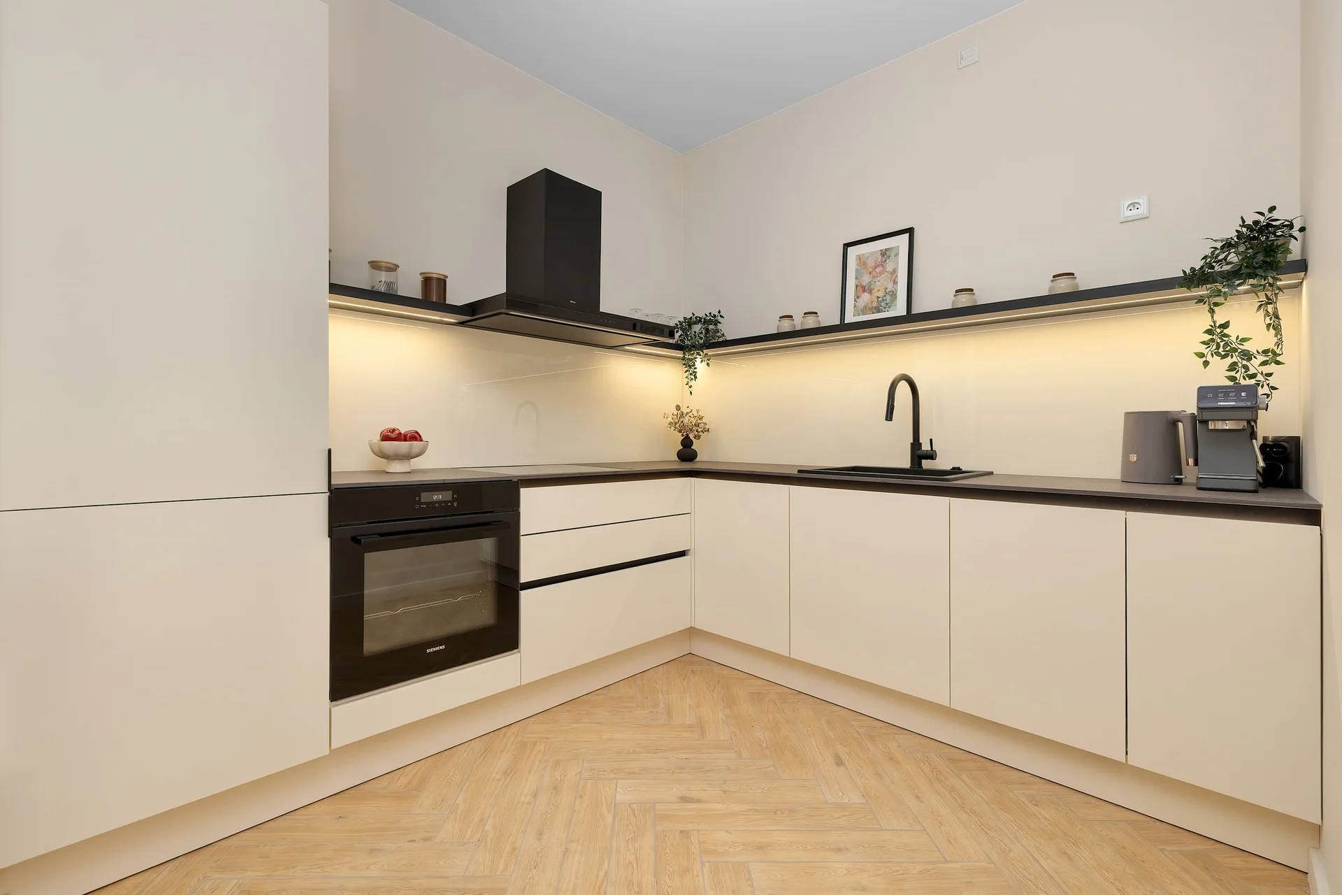 A sleek L-shaped kitchen combines white cabinetry with black countertops and integrated appliances. Minimalist finishes give the cooking area a clean, contemporary appearance.