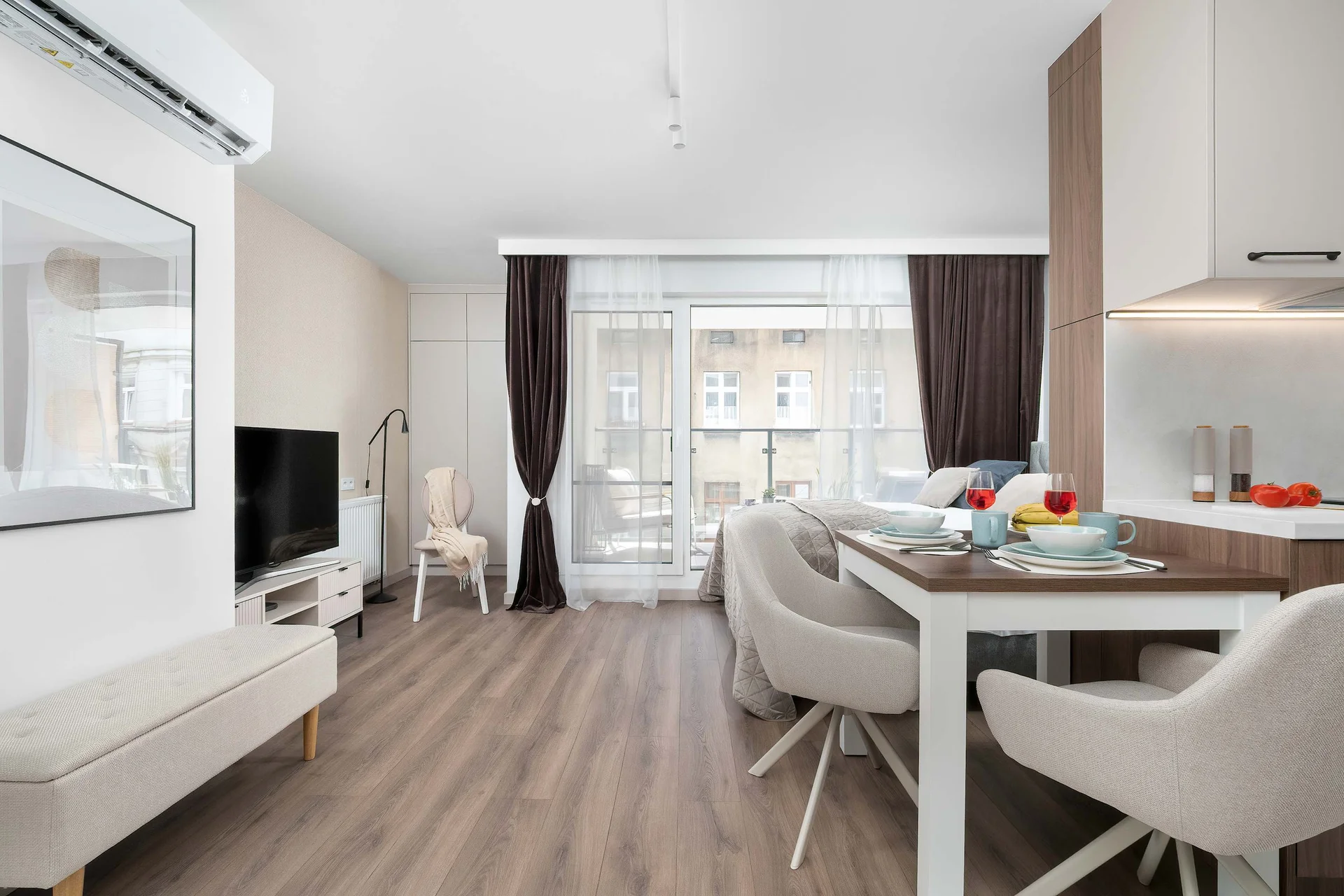 A bright studio view highlights a modern wooden dining table set with white chairs beneath cozy, warm lighting. The open floor plan extends toward a sleeping area, separated visually by clever layouts.