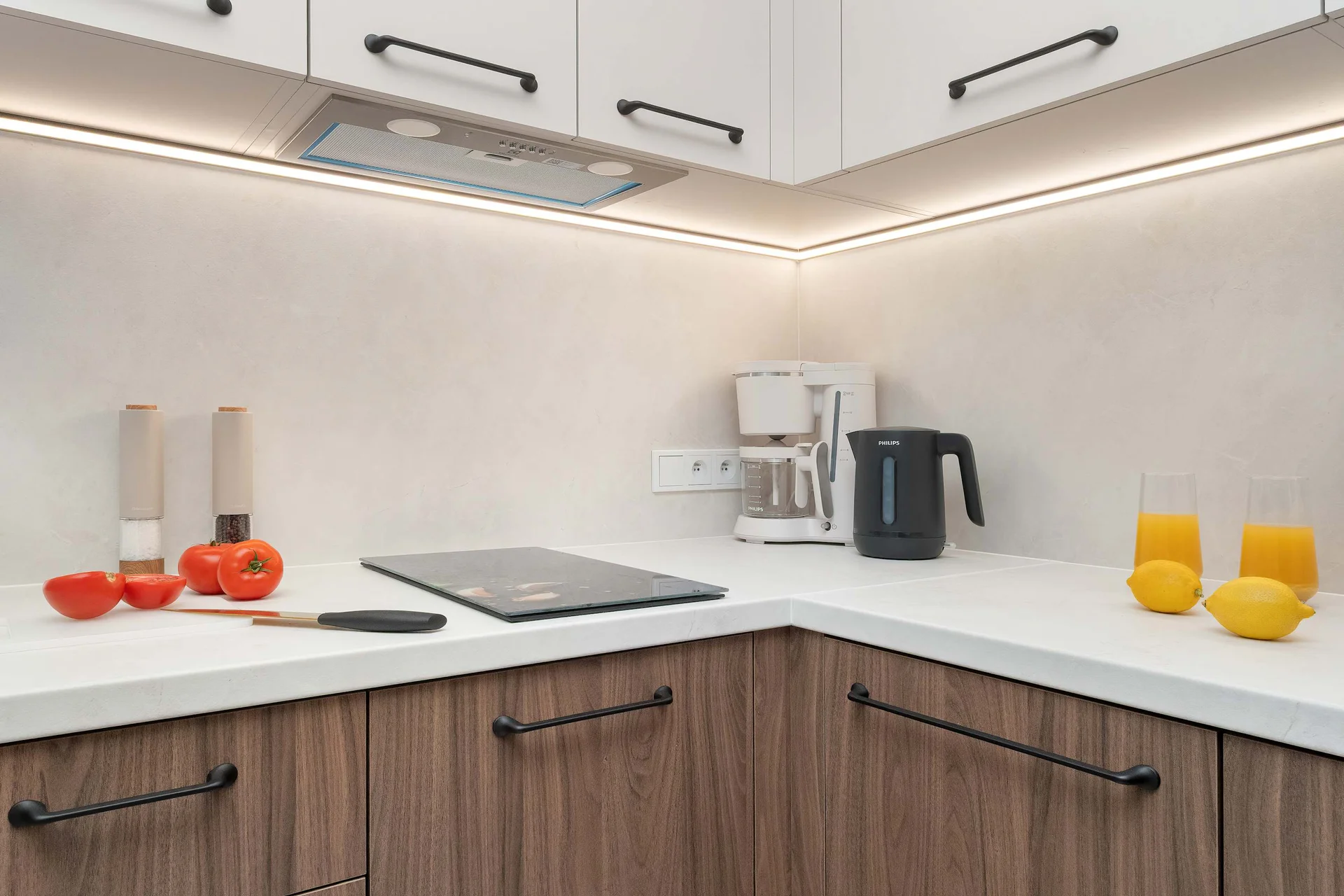 A compact kitchen corner features sleek white countertops and clean, light-colored upper cabinetry. Elegant wooden lower cabinets house integrated cooking appliances beneath bright LED strip lighting.