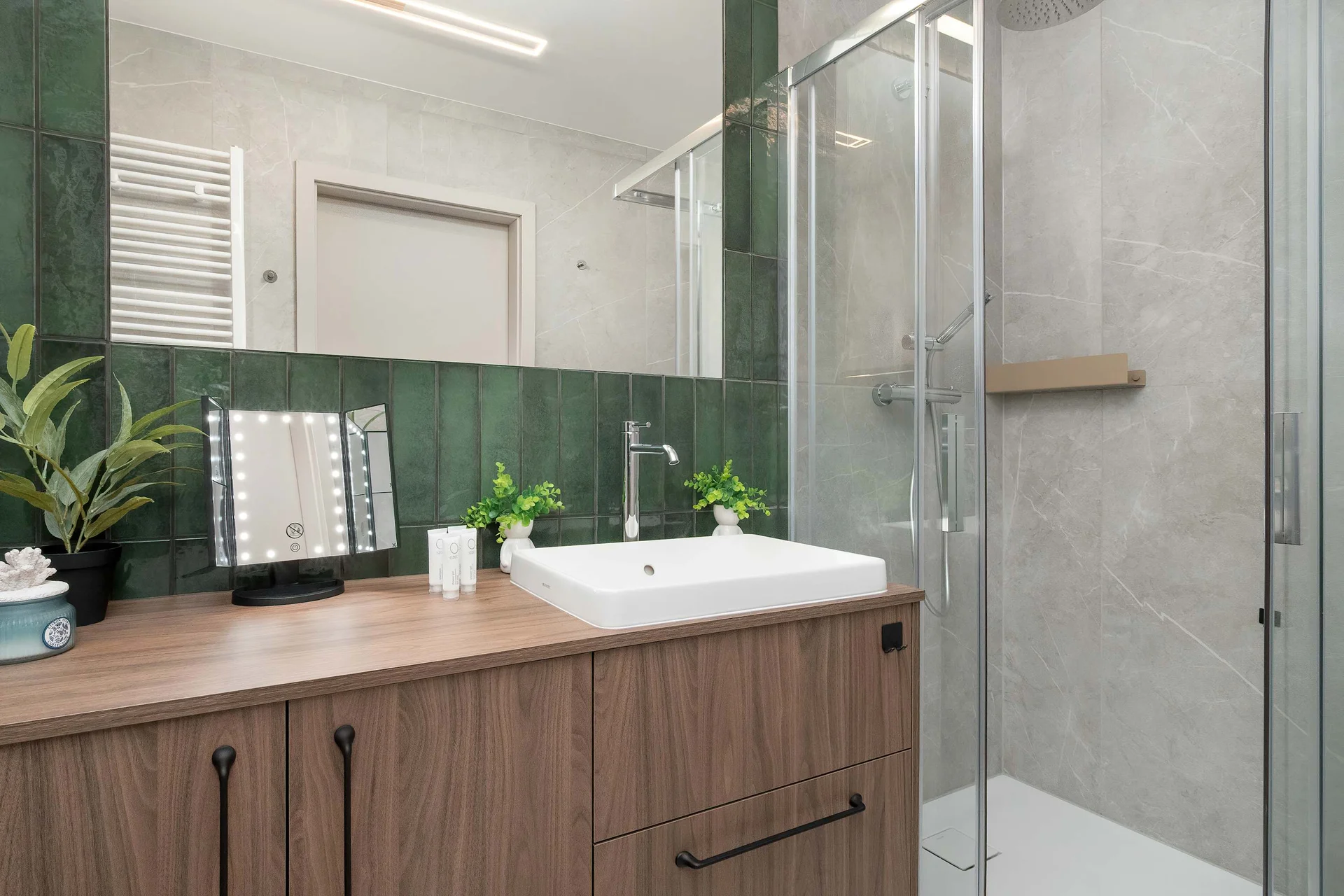 The functional bathroom features a striking olive-green tiled accent wall combined with warm wooden cabinetry. A clean white countertop washbasin with a modern chrome faucet sits below a large wall mirror.