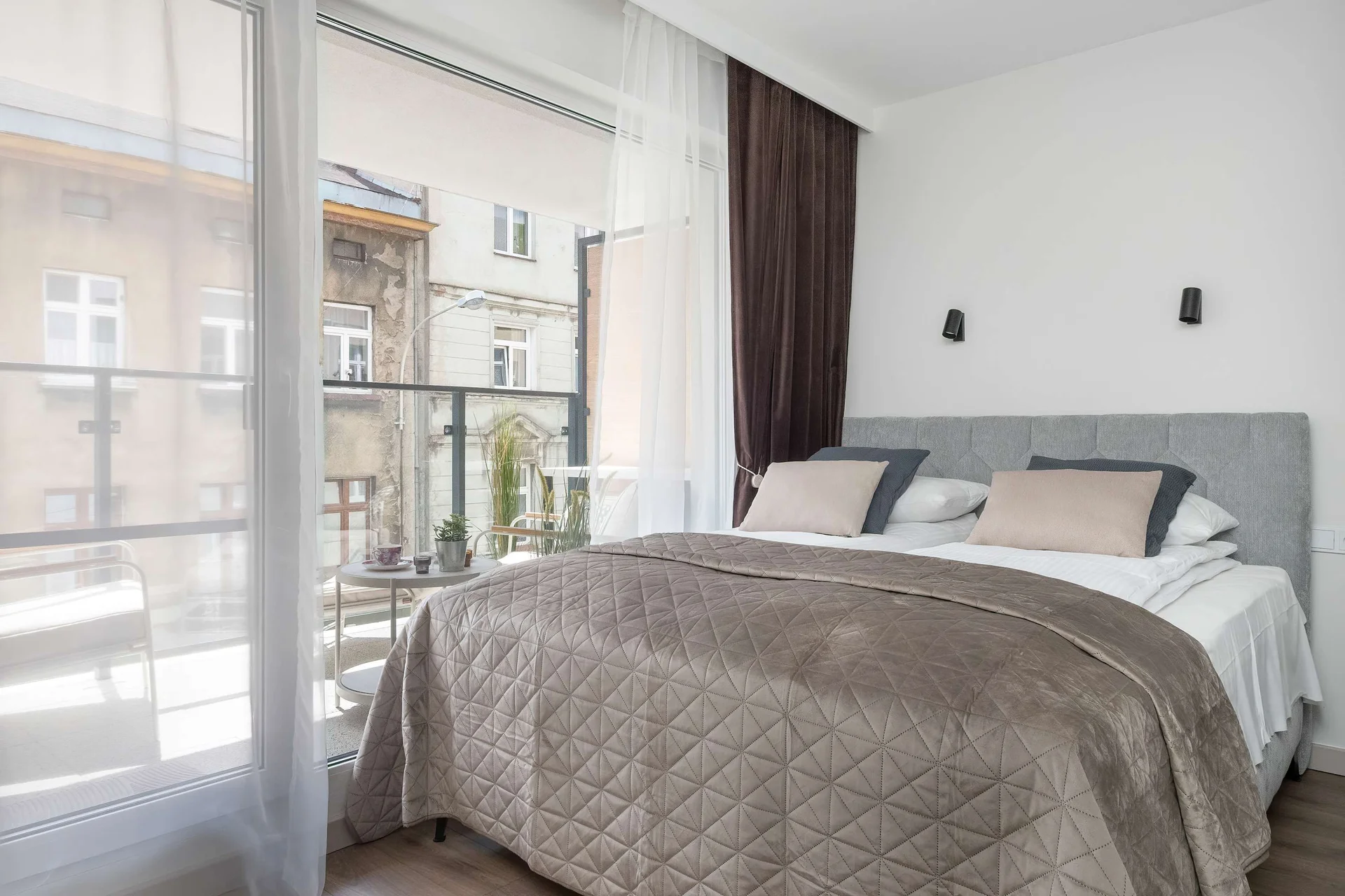 A beautifully dressed double bed is positioned next to a glass balcony door, allowing natural light to fill the room. Elegant grey and brown textiles cover the bed, creating a warm, inviting sleeping space.