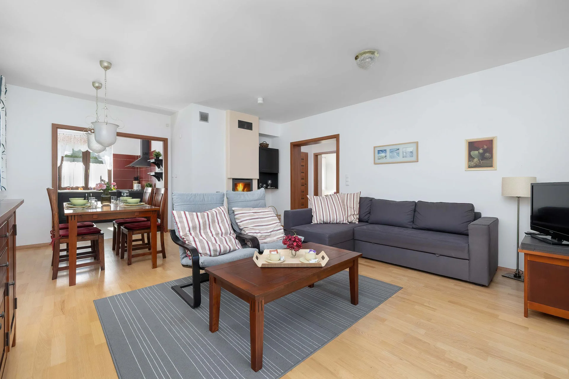 A bright living room with a gray sofa, coffee table, TV, and dining area. Large windows make the space feel open and inviting.