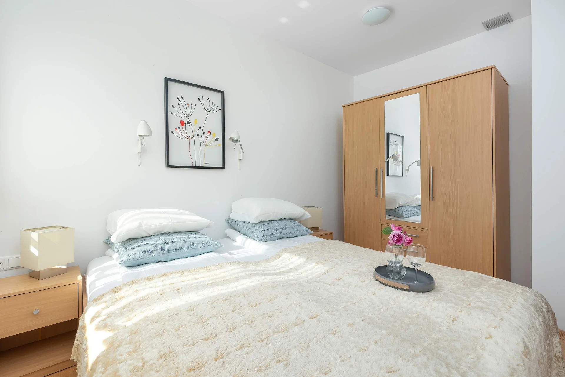 A cozy bedroom with a bed, wooden wardrobe, and bedside table. A window fills the room with natural light.