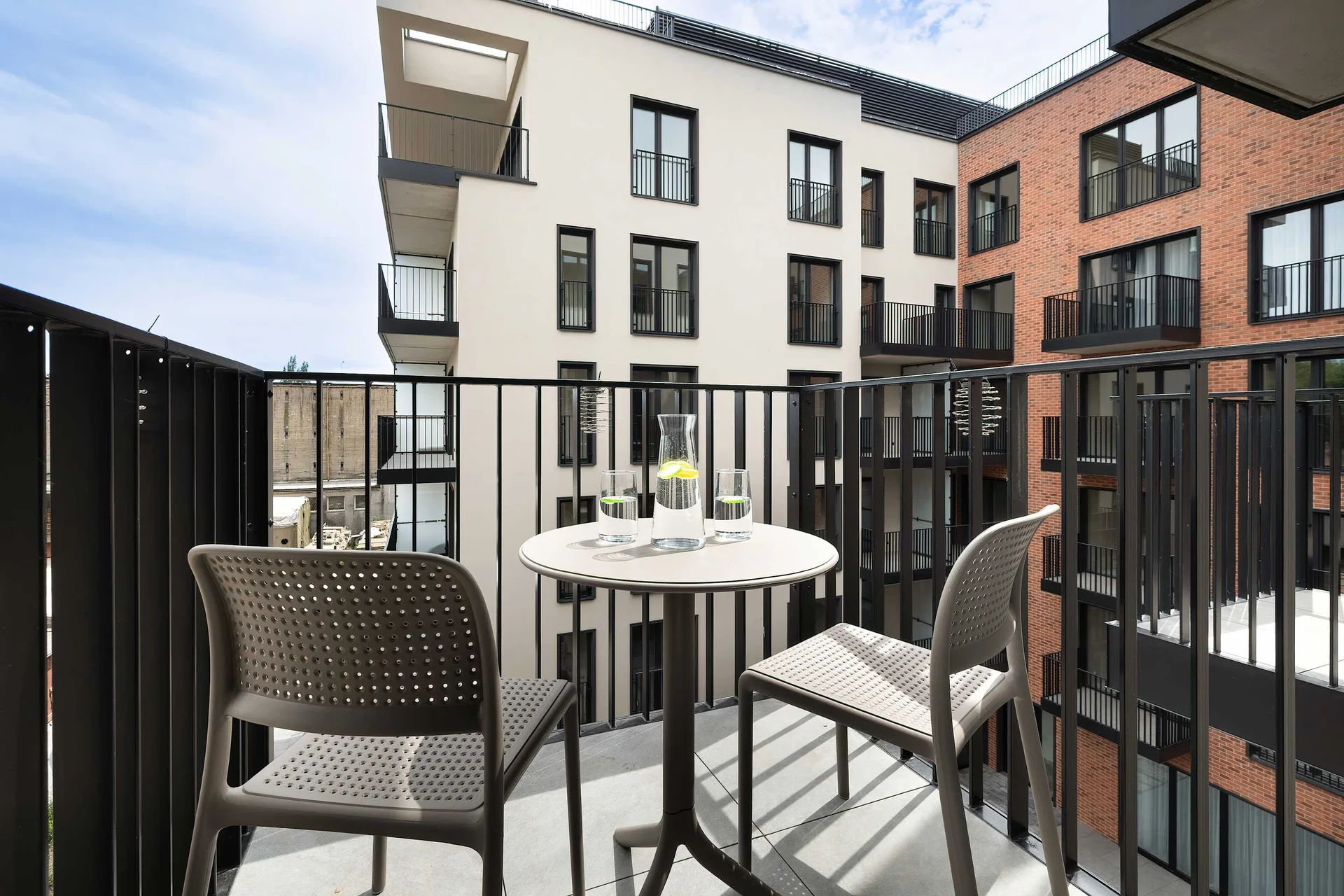 A private balcony is furnished with a table and chairs overlooking the courtyard and neighboring apartment buildings. The outdoor space is perfect for enjoying fresh air or a morning coffee.