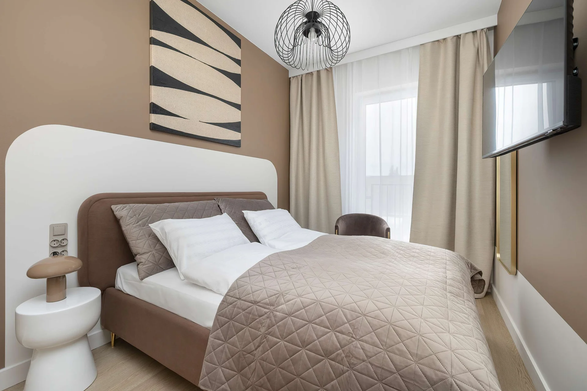 A stylish bedroom features a comfortable double bed with upholstered headboard, modern bedside tables, and a decorative accent wall. Soft lighting and neutral colors create a relaxing atmosphere.