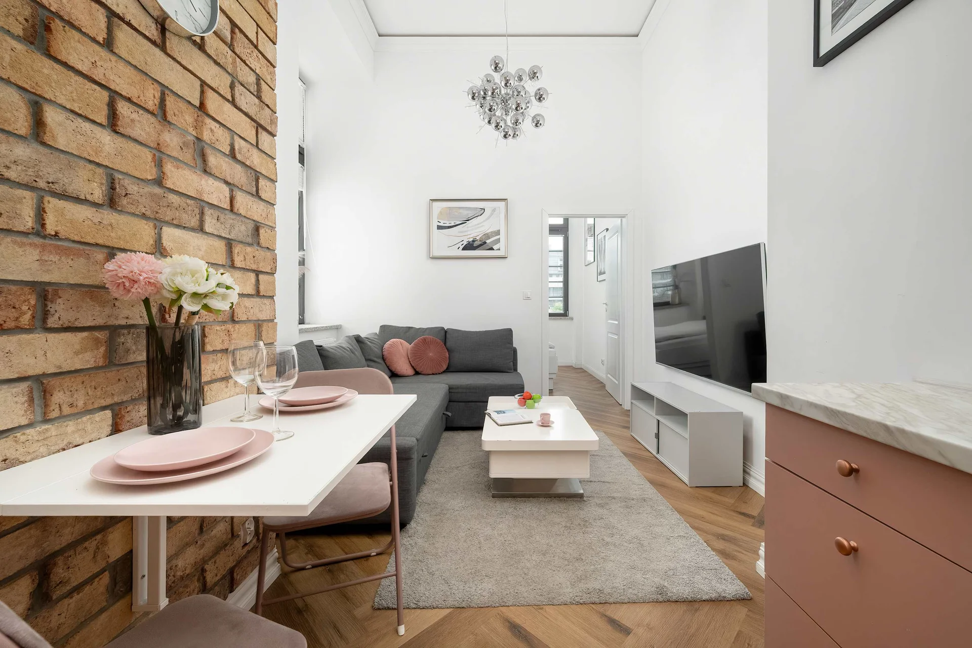 A bright living room with a comfortable sofa, a coffee table, and an exposed brick wall. The open layout connects the seating area with the kitchen.