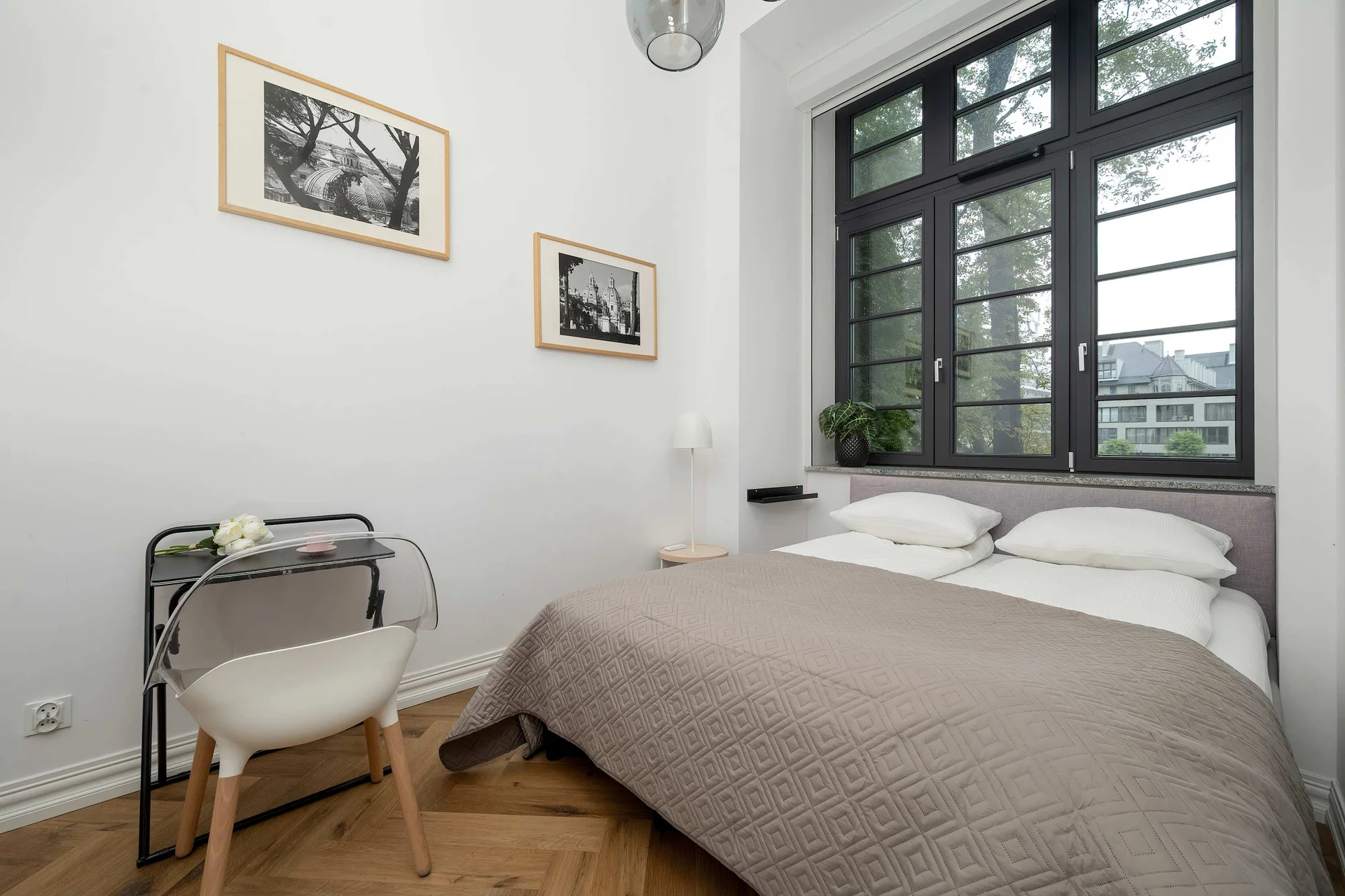 A cozy bedroom with a double bed, a desk, and large windows. The room is bright and simply decorated.
