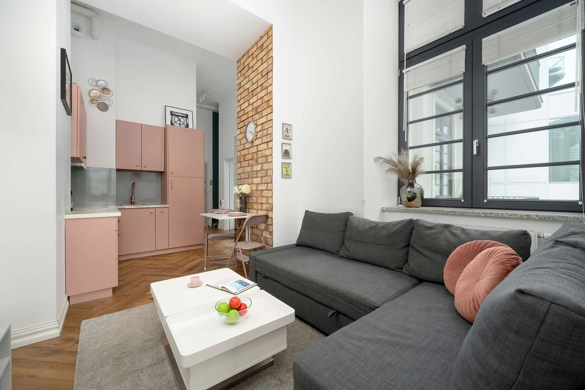 A stylish living room with a sofa, a coffee table, and large windows. The seating area is connected to a modern kitchenette.