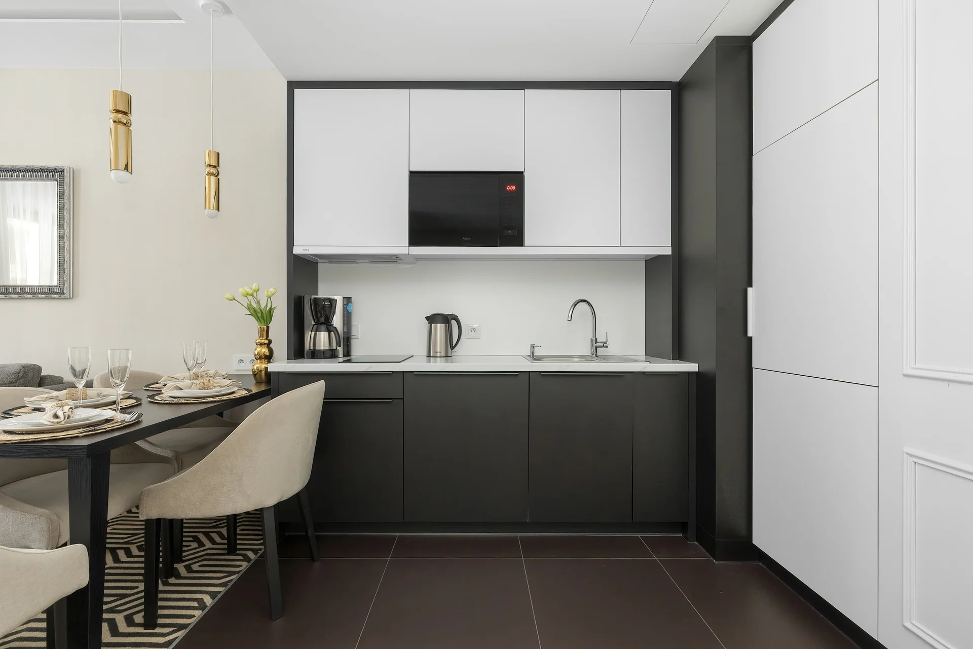 A modern kitchenette offers sleek black-and-white cabinetry, integrated appliances, and generous workspace. The minimalist design keeps the area bright and functional.