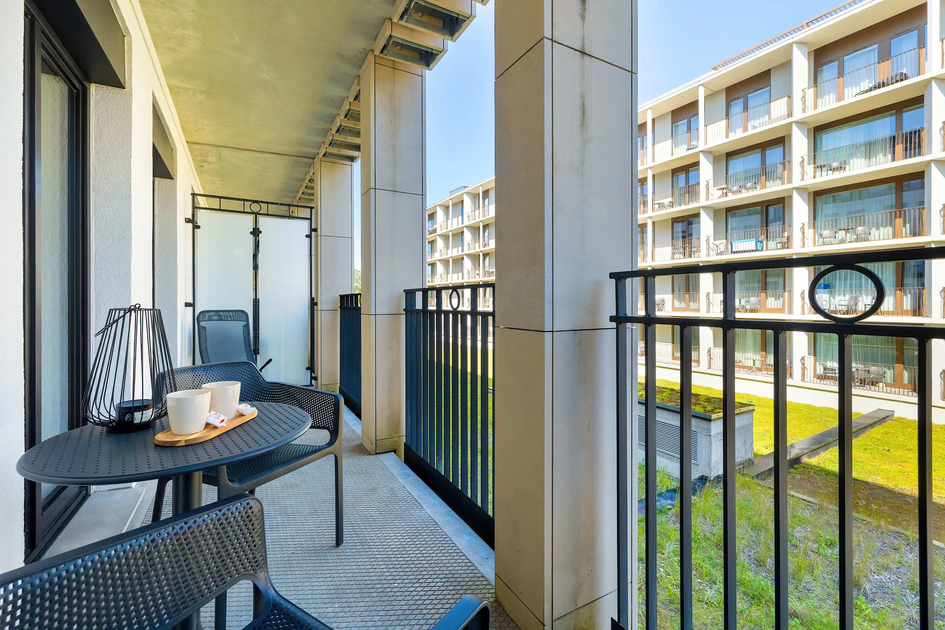 A covered balcony is furnished with a small table and chairs overlooking landscaped grounds and neighboring apartment buildings. The outdoor space is ideal for relaxing with a drink.