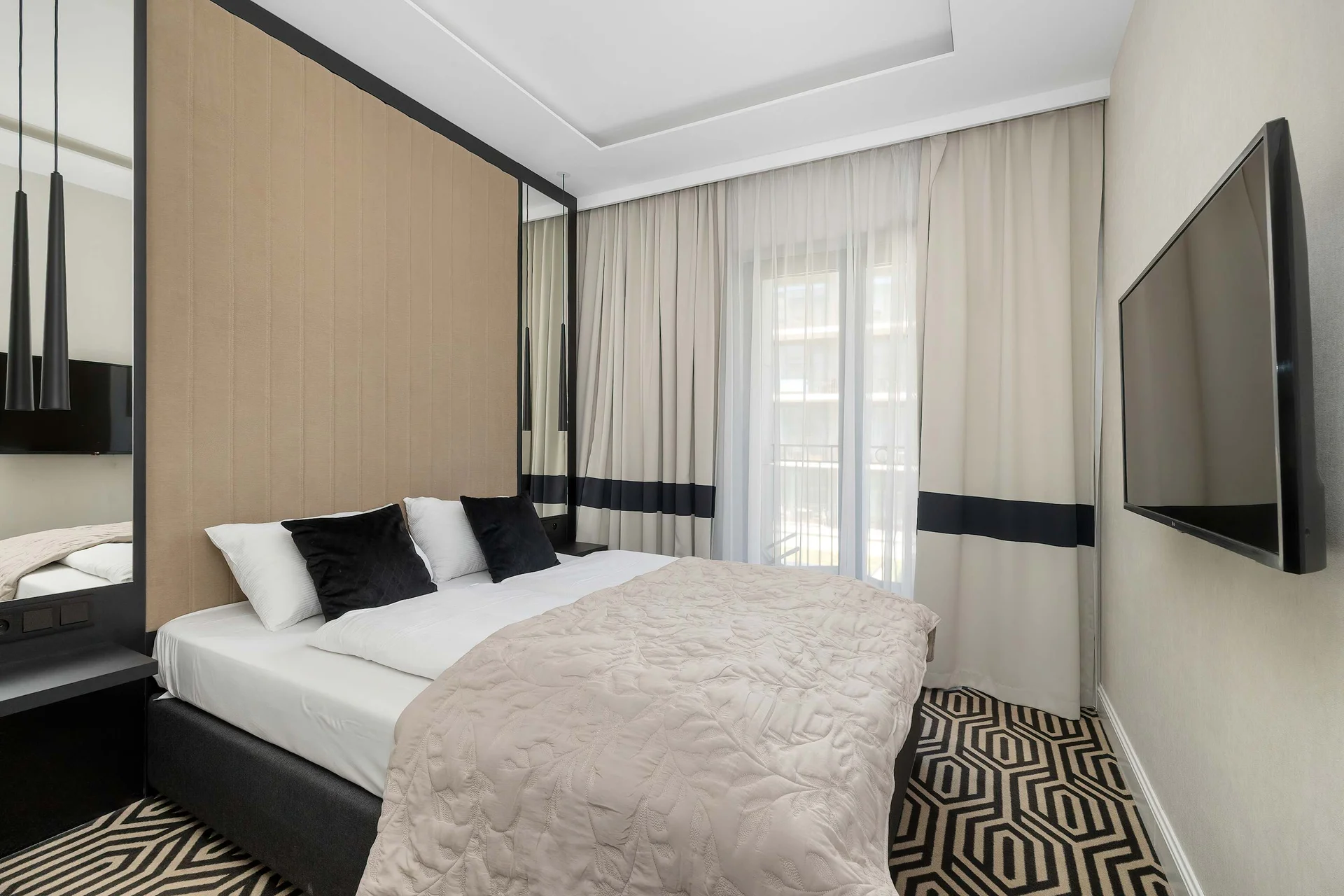 A sophisticated bedroom features a plush double bed, elegant wall paneling, and a wall-mounted TV opposite the bed. Soft curtains and warm tones create a peaceful atmosphere.