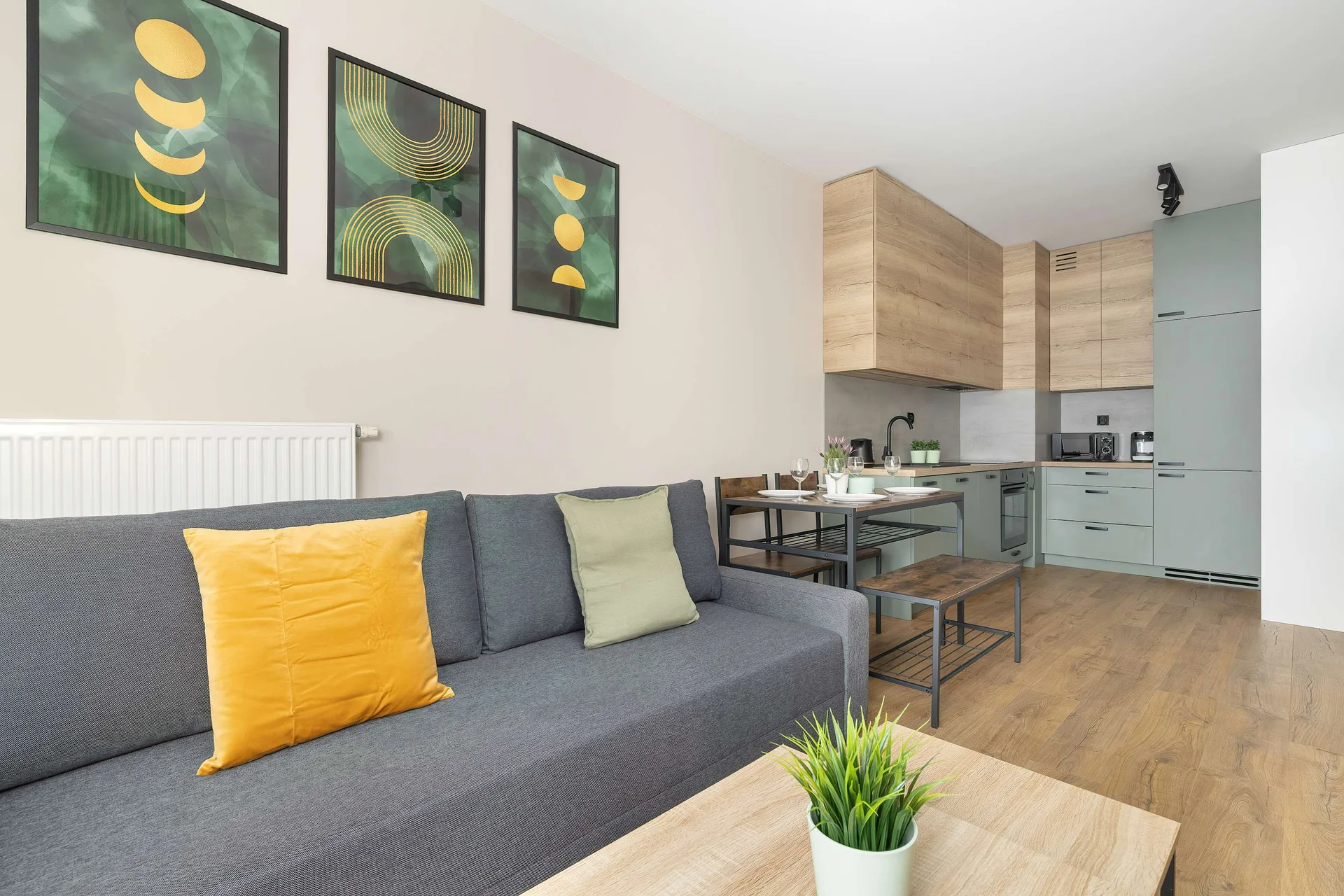 A bright open-plan living space combines the grey sofa with a view towards the adjacent kitchen area. Three matching botanical prints adorn the wall, and a wooden coffee table sits in the foreground on the light laminate flooring.