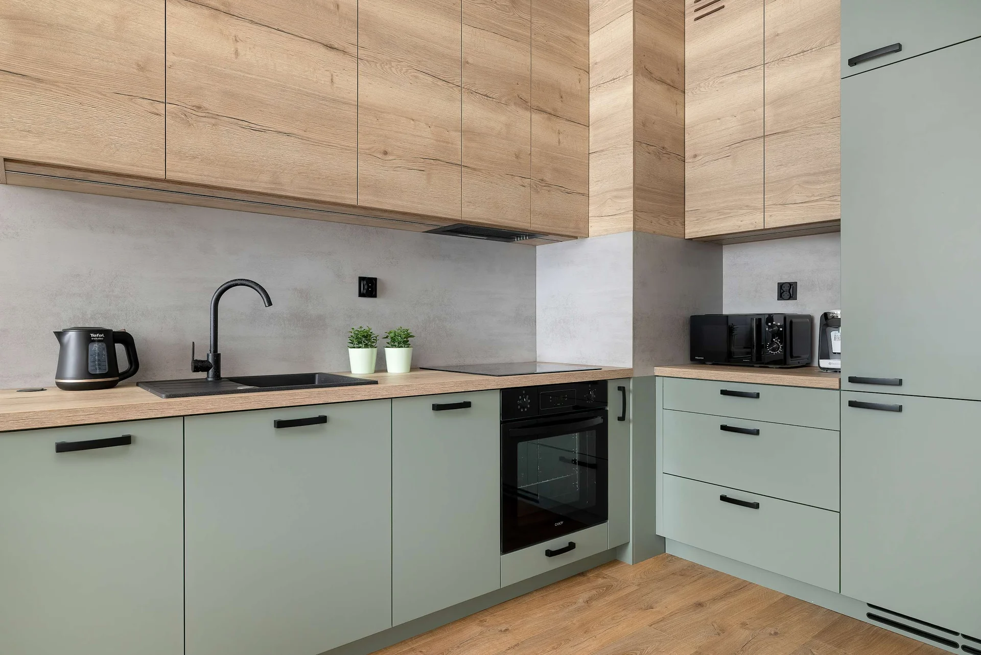 A modern kitchen corner showcases sage green lower cabinets paired with light wood upper cabinetry and a matching countertop. A black sink, sleek faucet, and built-in black oven are integrated into the seamless design.