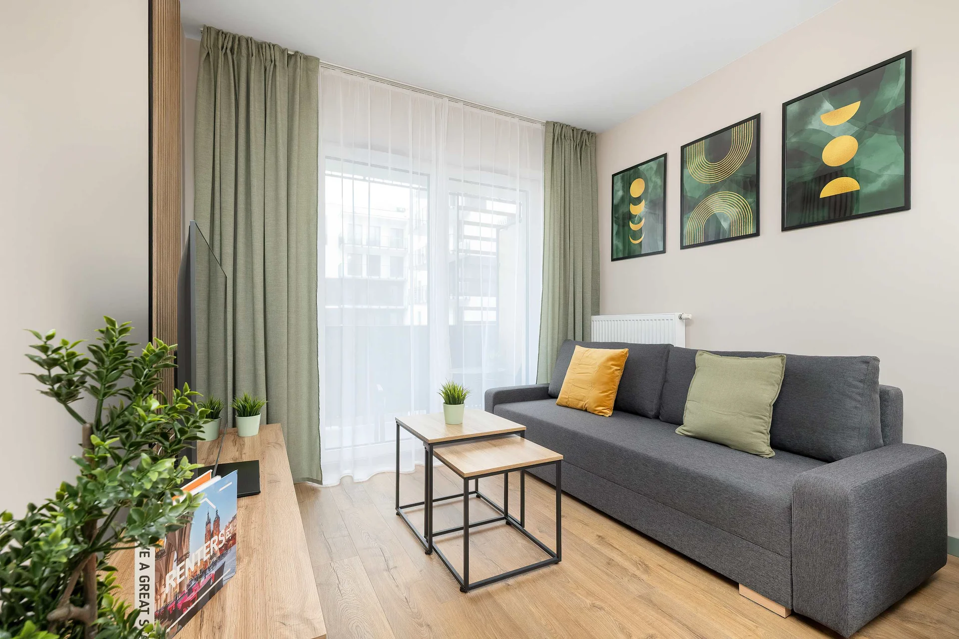 A cozy living room features a modern grey sofa decorated with green and yellow cushions. A small black-framed coffee table sits on the light wood floor, while three green botanical art prints hang on the wall above the couch.