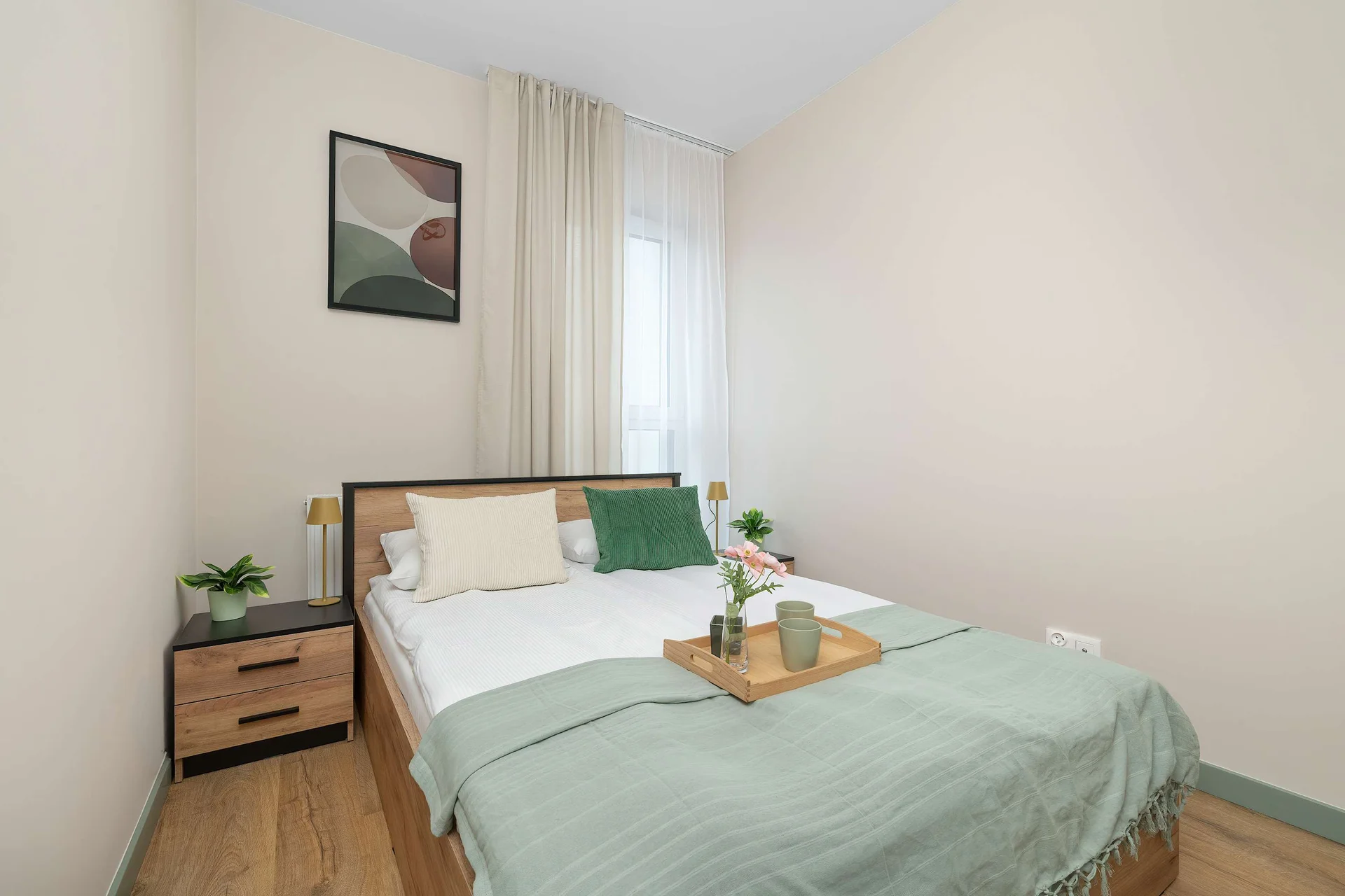 A minimalist bedroom displays a comfortable double bed made up with white linens and a soft green throw blanket. A wooden tray with small decorative items rests on the bed.