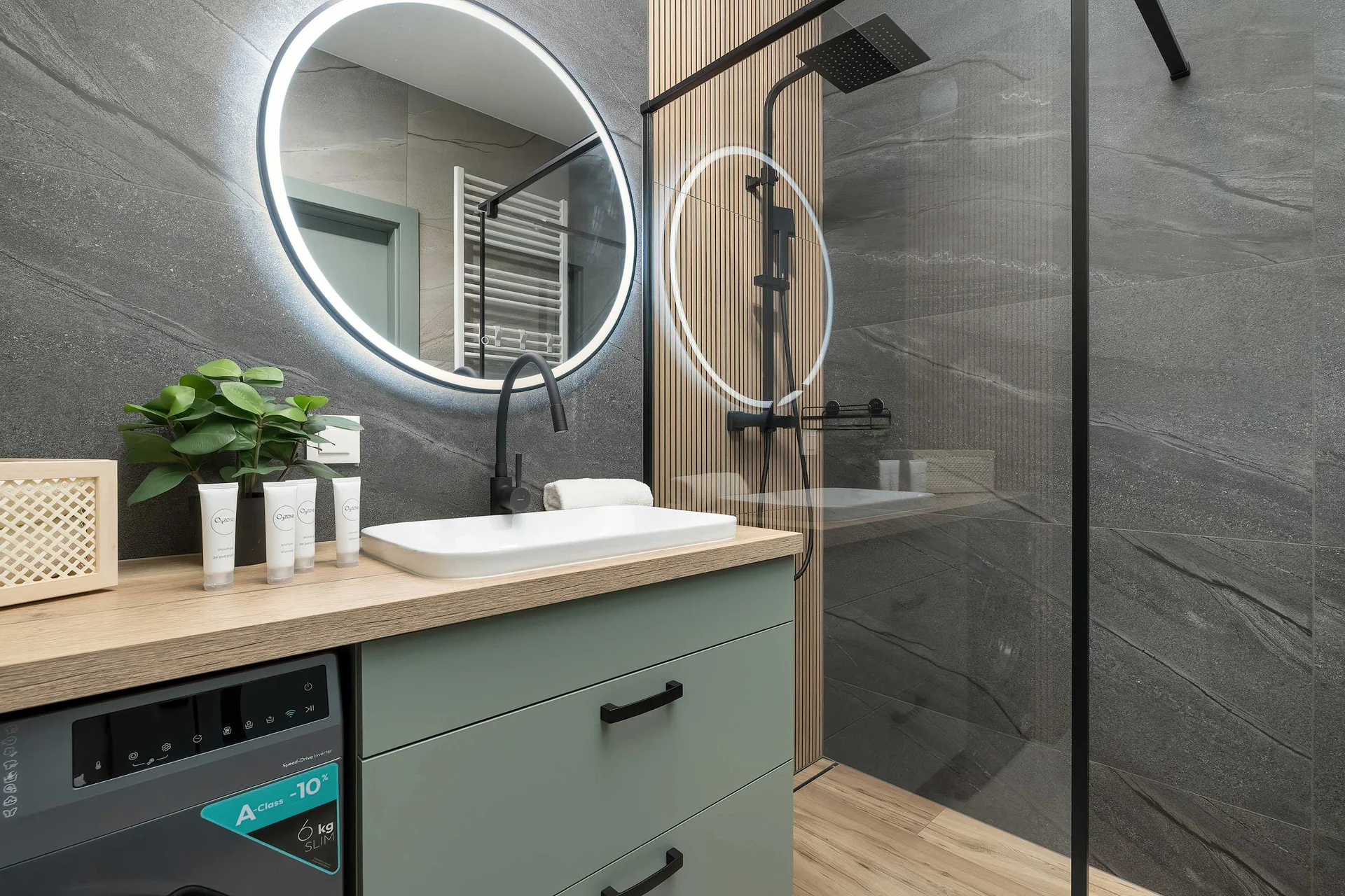 A stylish bathroom setup highlights a round LED-backlit mirror mounted on a dark textured slate wall. A white vessel sink rests on a light wood countertop above a sage green vanity drawer next to a glass shower screen.