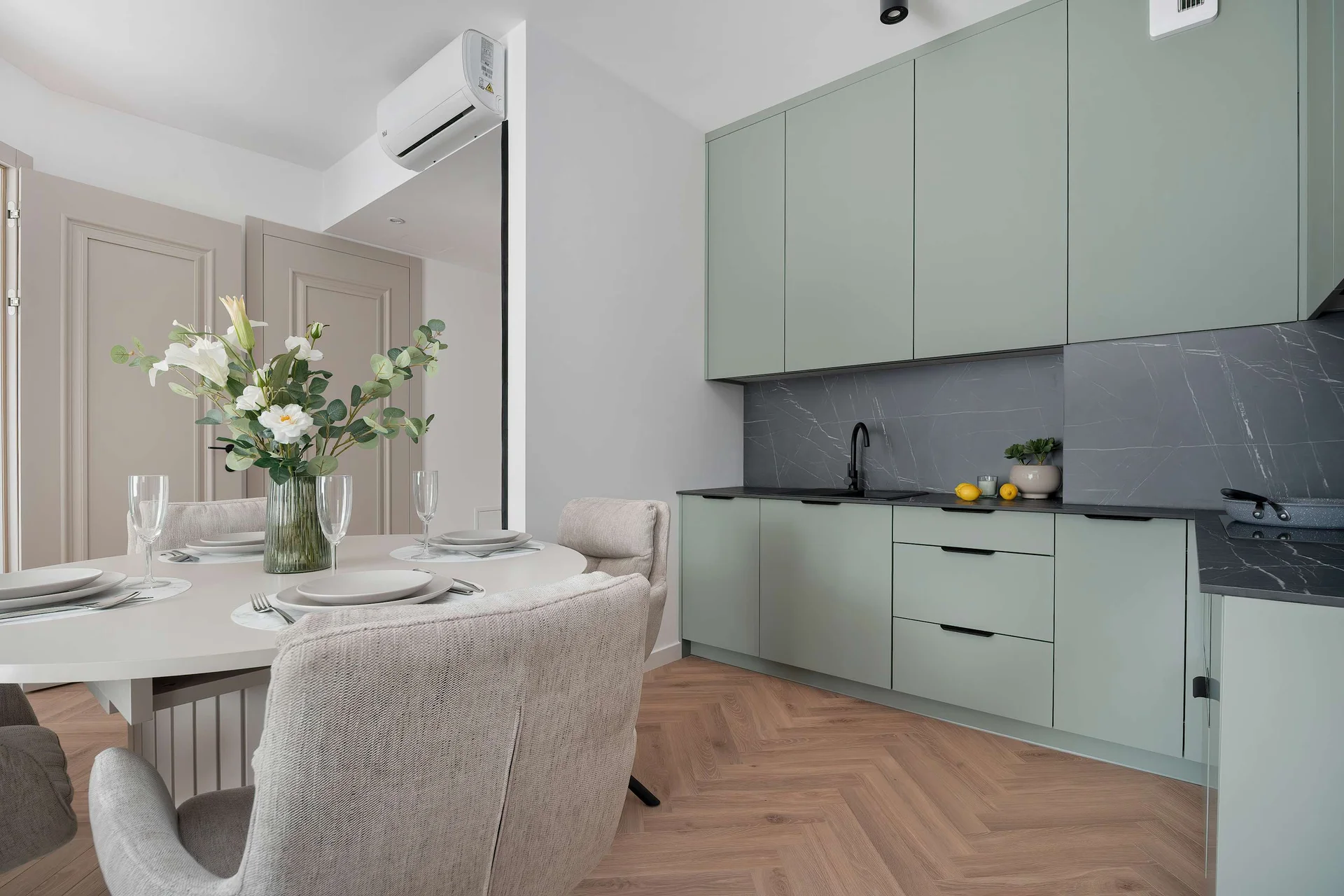 A sleek sage-green kitchen displays modern handleless cabinets and dark marble-patterned countertops. A round dining table sits in the foreground.
