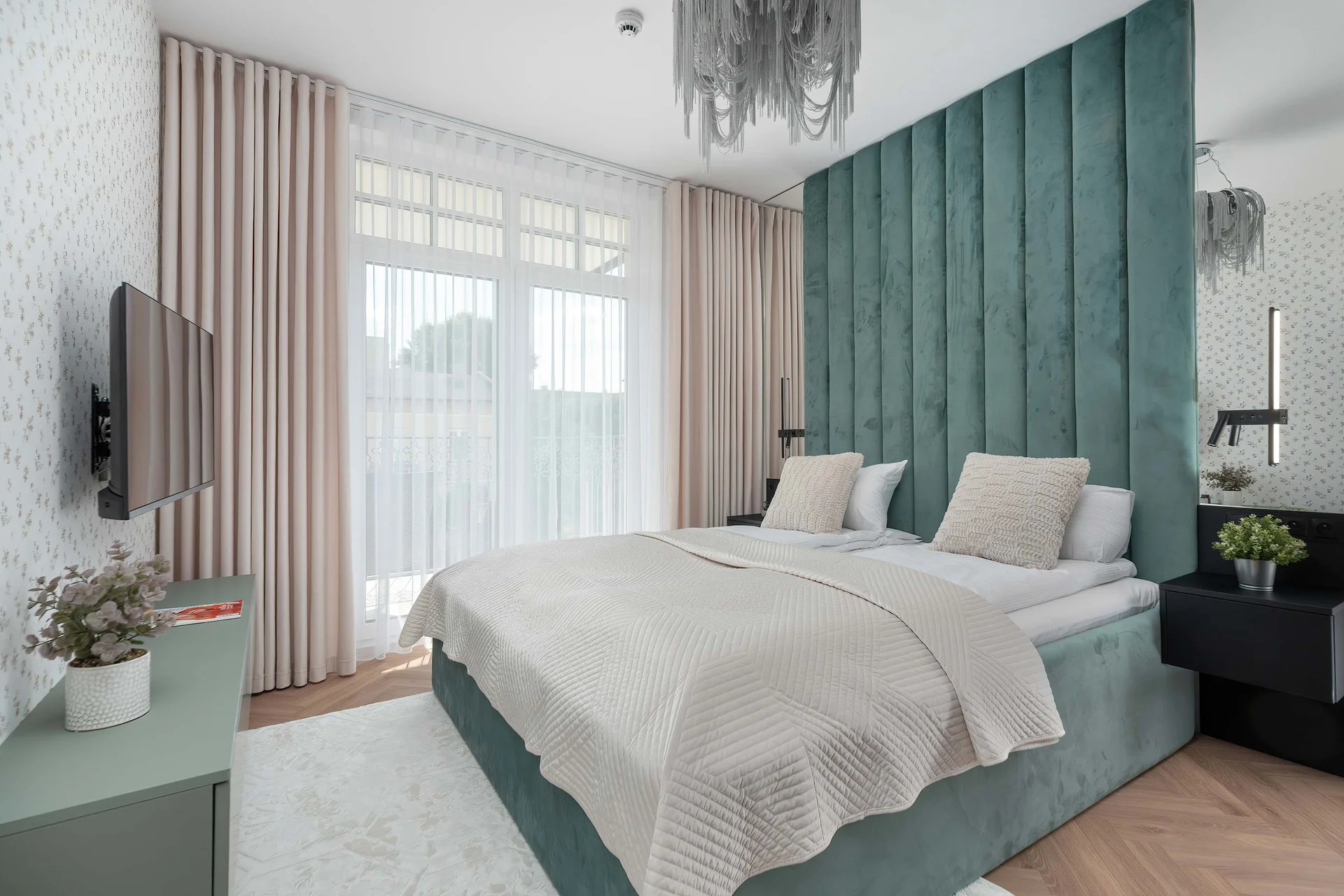 A bedroom features a double bed with a teal velvet channel-tufted headboard. Sheer white curtains and soft pink drapes frame the large balcony window.