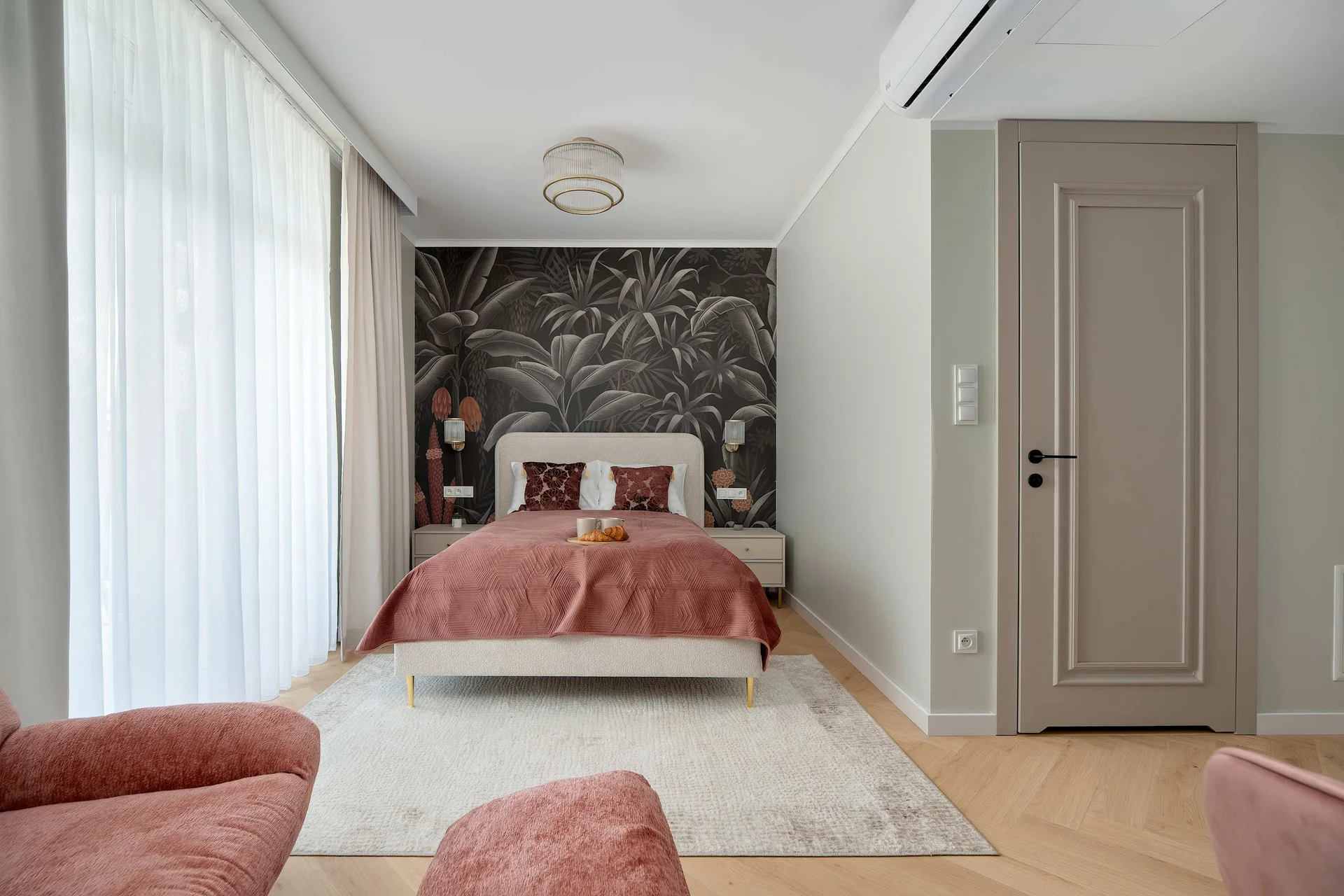 A bright bedroom with a double bed, pink armchairs, and a decorative accent wall. Large windows provide plenty of natural light.