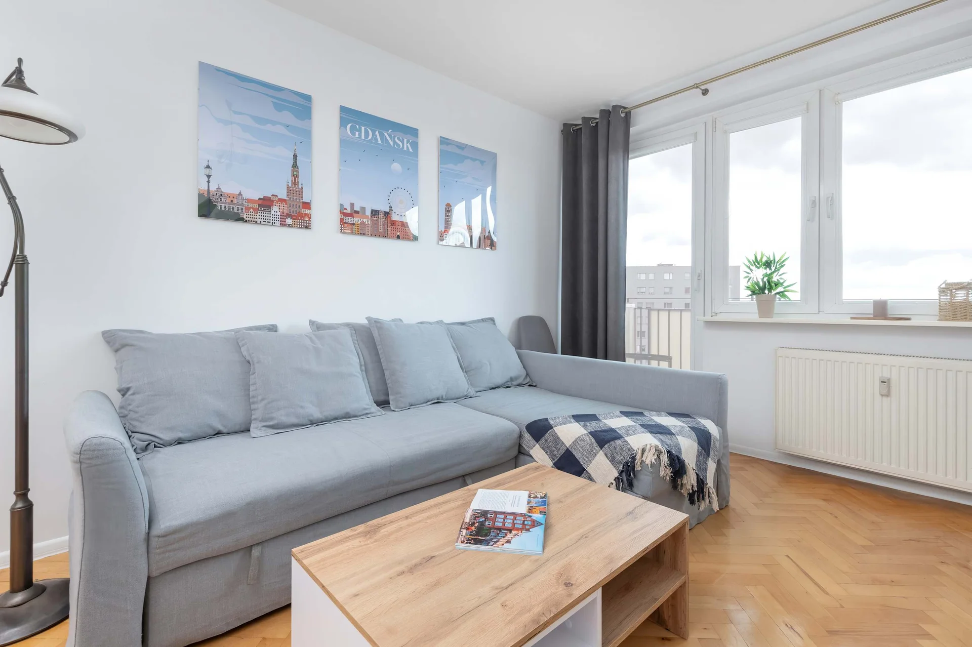 Bright living room with a comfortable grey sofa, coffee table, TV, and large windows providing plenty of natural light.