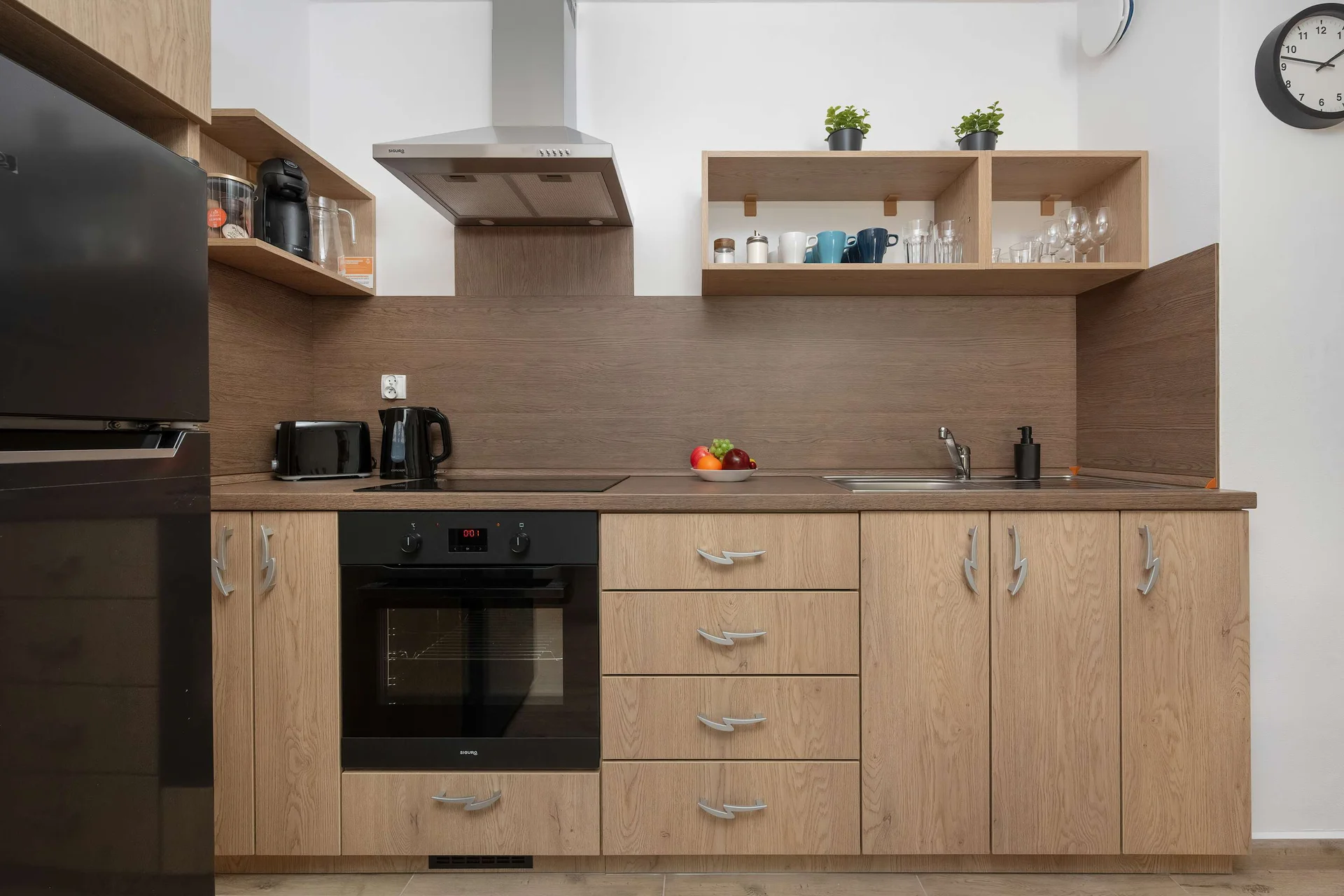 A modern kitchenette features warm wooden cabinetry and a stylish black backsplash. An oven and refrigerator are seamlessly integrated into the functional cooking space.