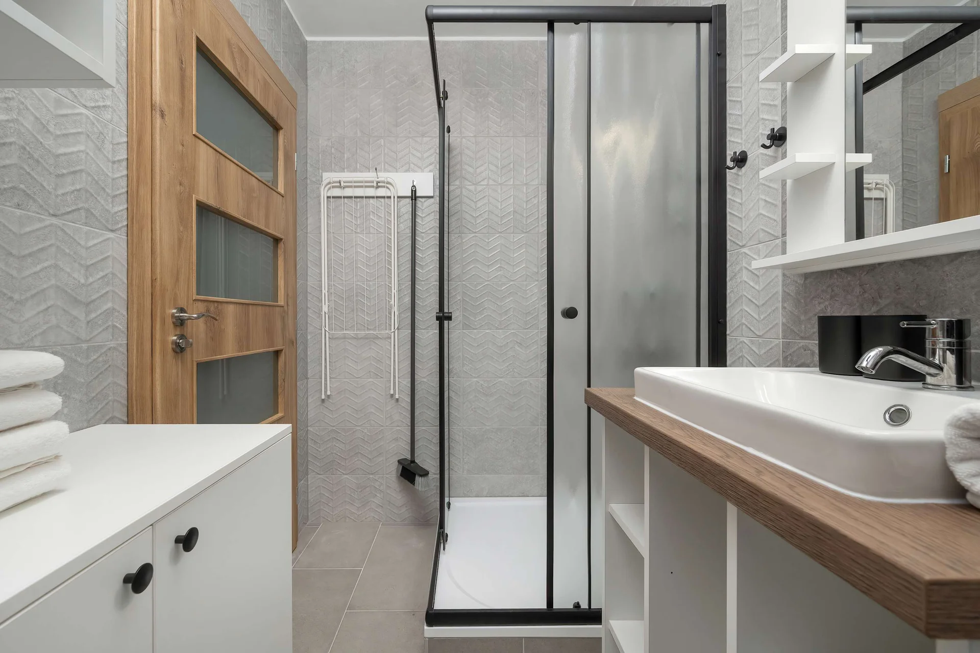 A clean, modern bathroom includes a glass shower cabin and light grey tiling. A white rectangular sink sits elegantly on a floating wooden countertop.
