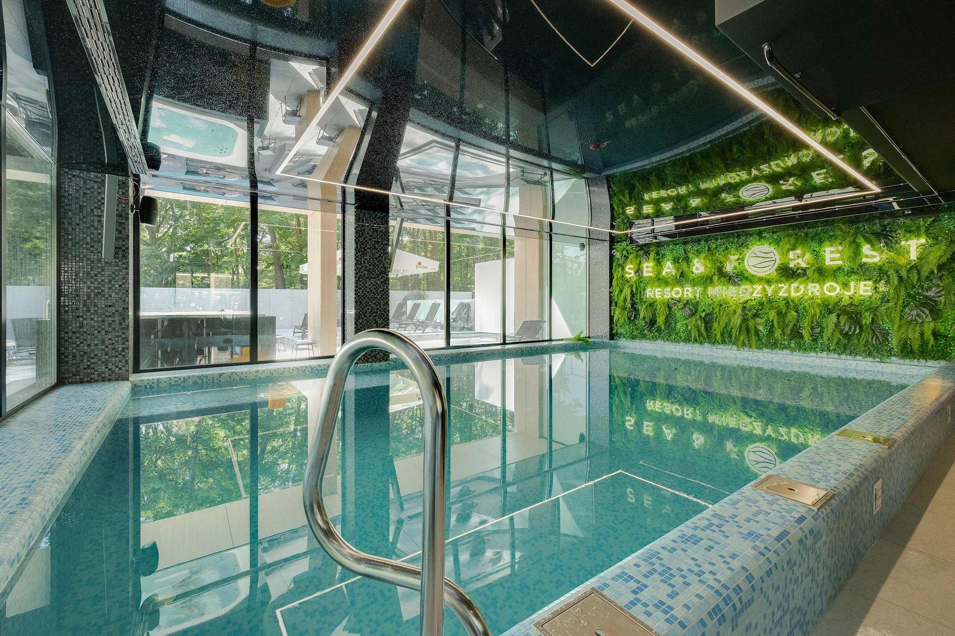 Modern indoor swimming pool with large glass windows, turquoise water, wooden details and lush green plants creating a relaxing atmosphere.