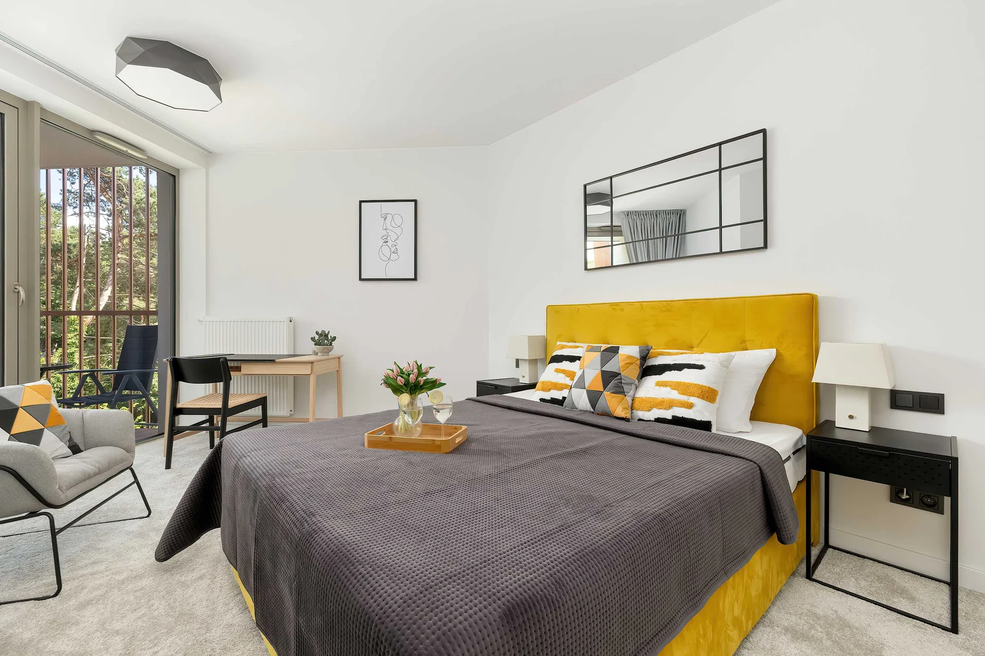 The bedroom displays a double bed with a yellow tufted headboard and dark grey bedding. A multi-pane mirror hangs above the headboard on a white wall.