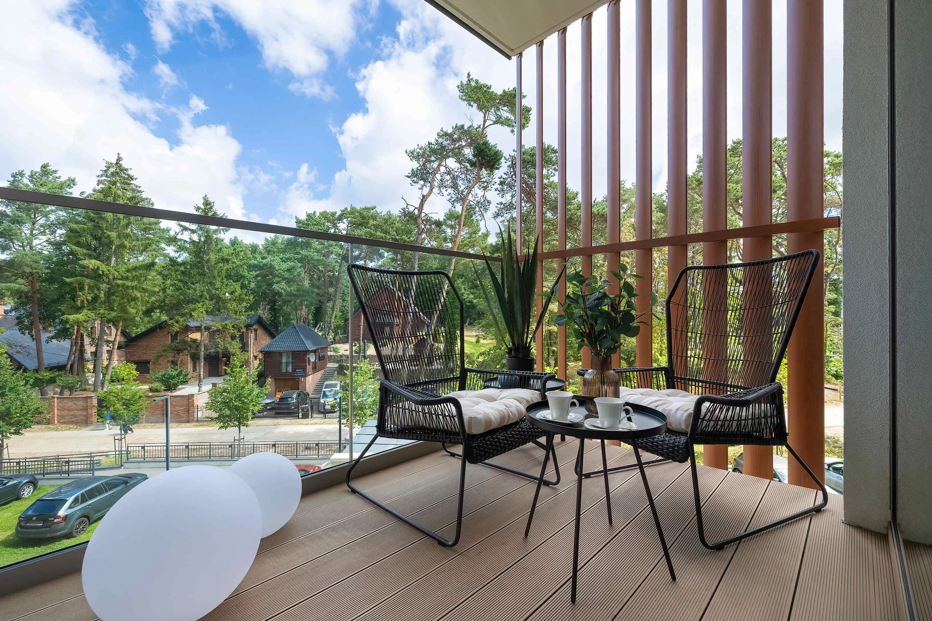 A covered wooden balcony with slatted privacy screens is furnished with two black chairs and a side table. The view overlooks lush green trees.