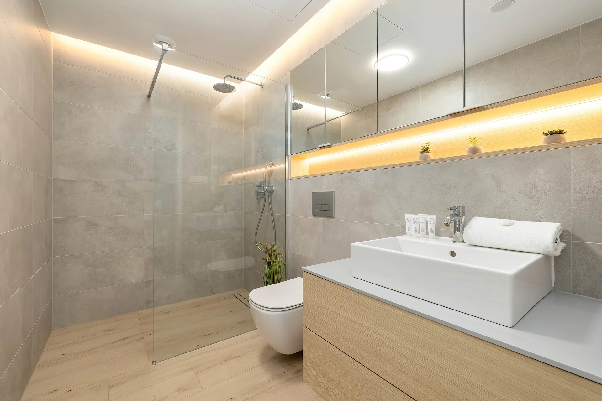 A spacious bathroom features large grey tiles, a walk-in shower with LED strip lighting, and a modern white vanity unit with a countertop washbasin.