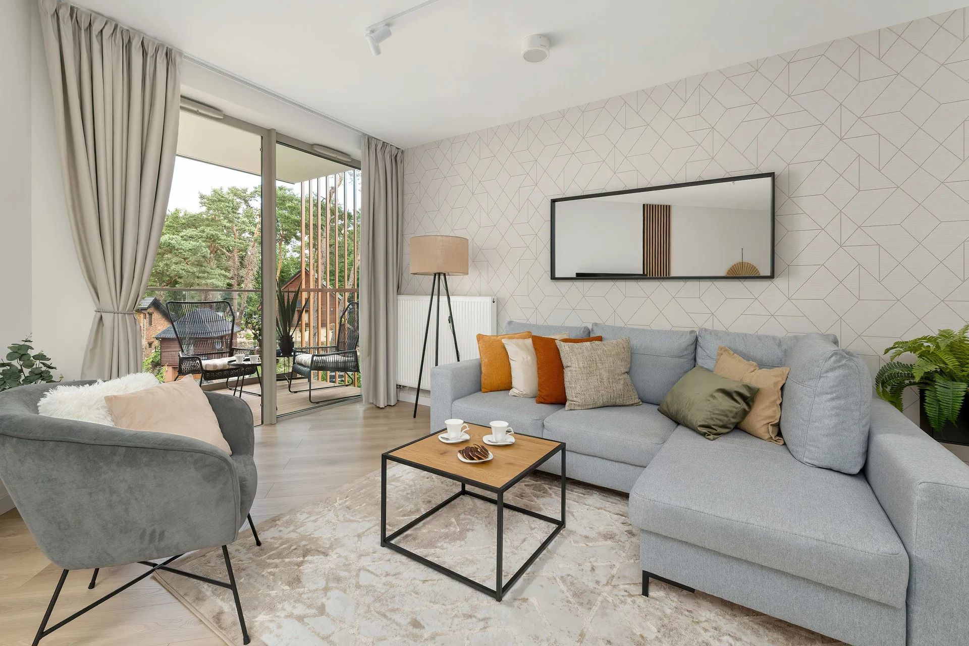 A bright living room features a light grey corner sofa styled with mustard throw pillows, a glass coffee table, and an armchair. Sliding glass doors lead onto a balcony.