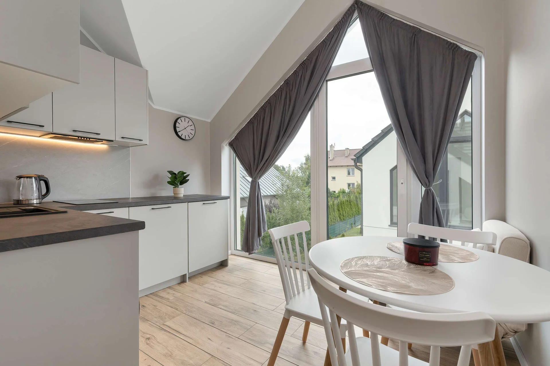 A dining area features a round wooden table and white chairs placed alongside tall floor-to-ceiling windows. Flowing curtains and sloped architectural lines add a sophisticated touch to everyday meal times.