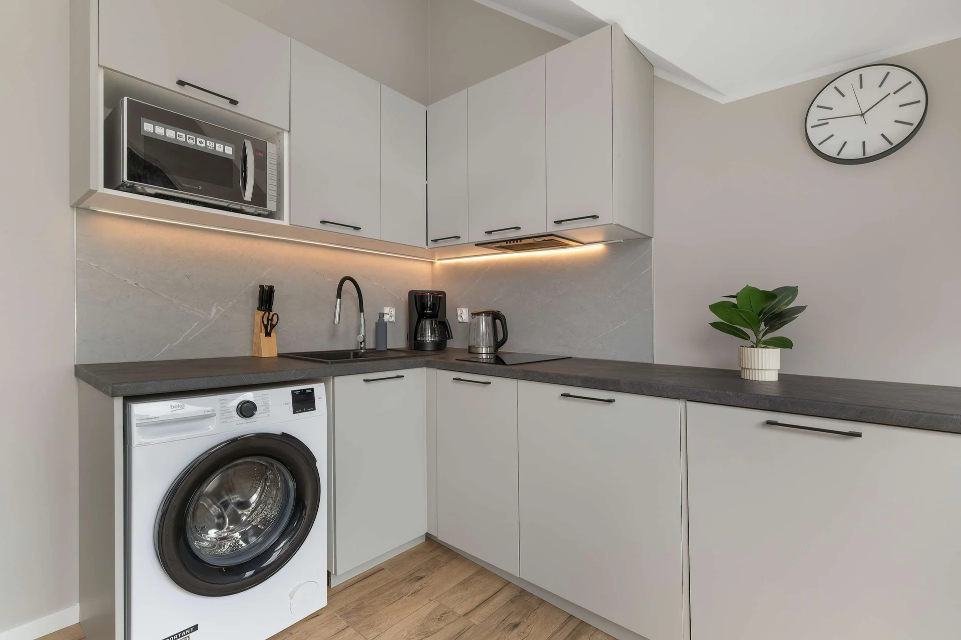 A practical kitchenette counter integrates an under-counter washing machine and a built-in microwave oven. Clean white tiling and a small decorative wall clock complete this efficient self-catering setup.