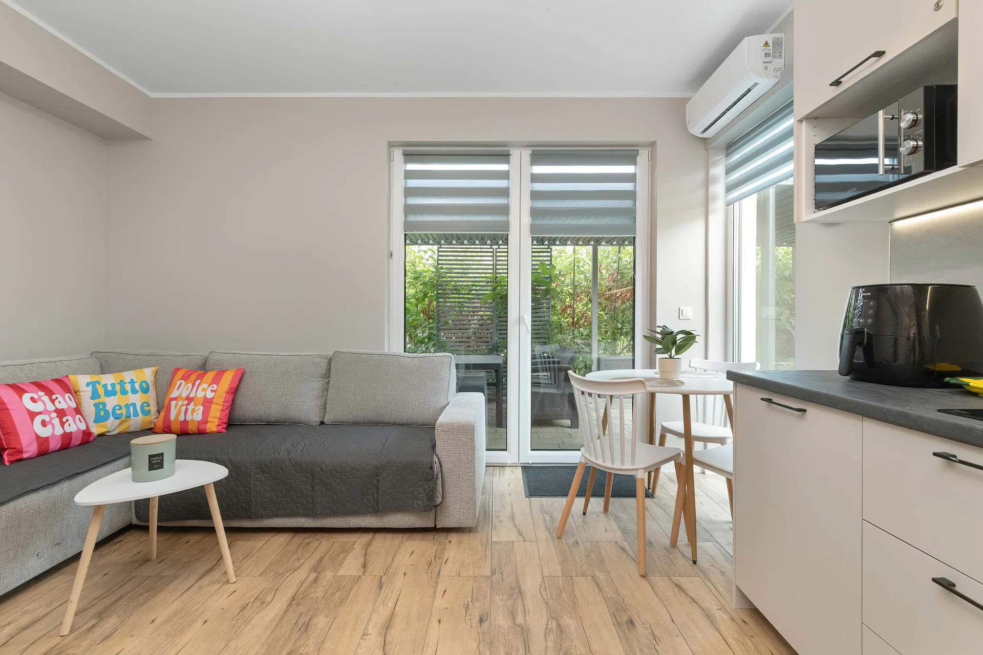 An open living room features a dark grey corner sofa with colorful cushions and light wood floors. Glass doors lead directly out to a sunlit paved terrace. A clean kitchenette lines the adjacent side of the space.