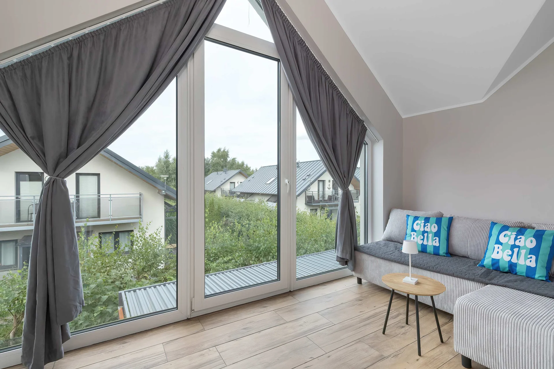 An airy upper-floor lounge boasts vaulted ceilings and floor-to-ceiling windows framed by dark curtains. A comfortable grey sofa with patterned cushions creates a serene setting for reading. Natural light fills the entire space.