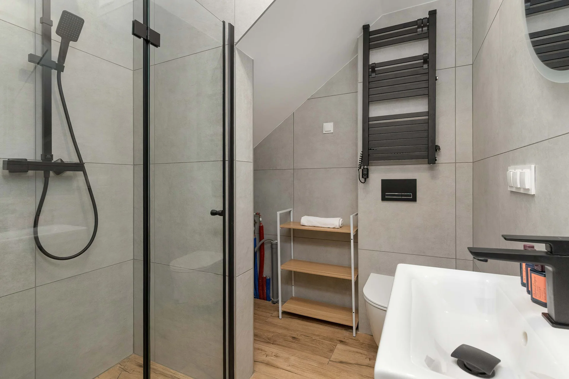 A modern bathroom is designed with grey ceramic wall tiles, a walk-in shower cubicle, and an oval vanity mirror. A black wall-hung sink faucet pairs cleanly with open wooden storage shelves fitted under the sloping ceiling.