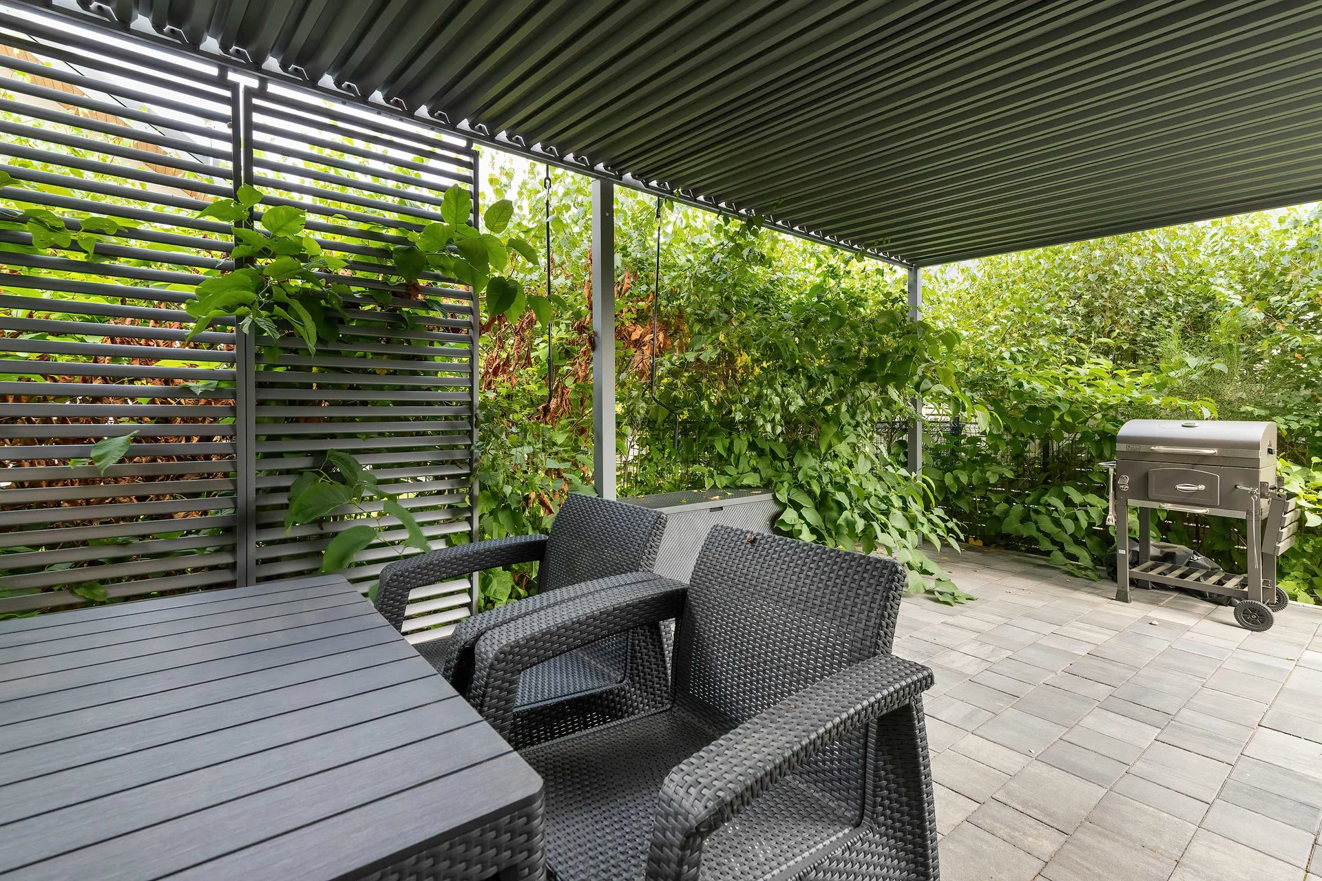 A private outdoor patio under a dark pergola is furnished with garden armchairs and a dining table. Vertical wooden slats draped in lush climbing vines create a sheltered, relaxing corner for alfresco meals.