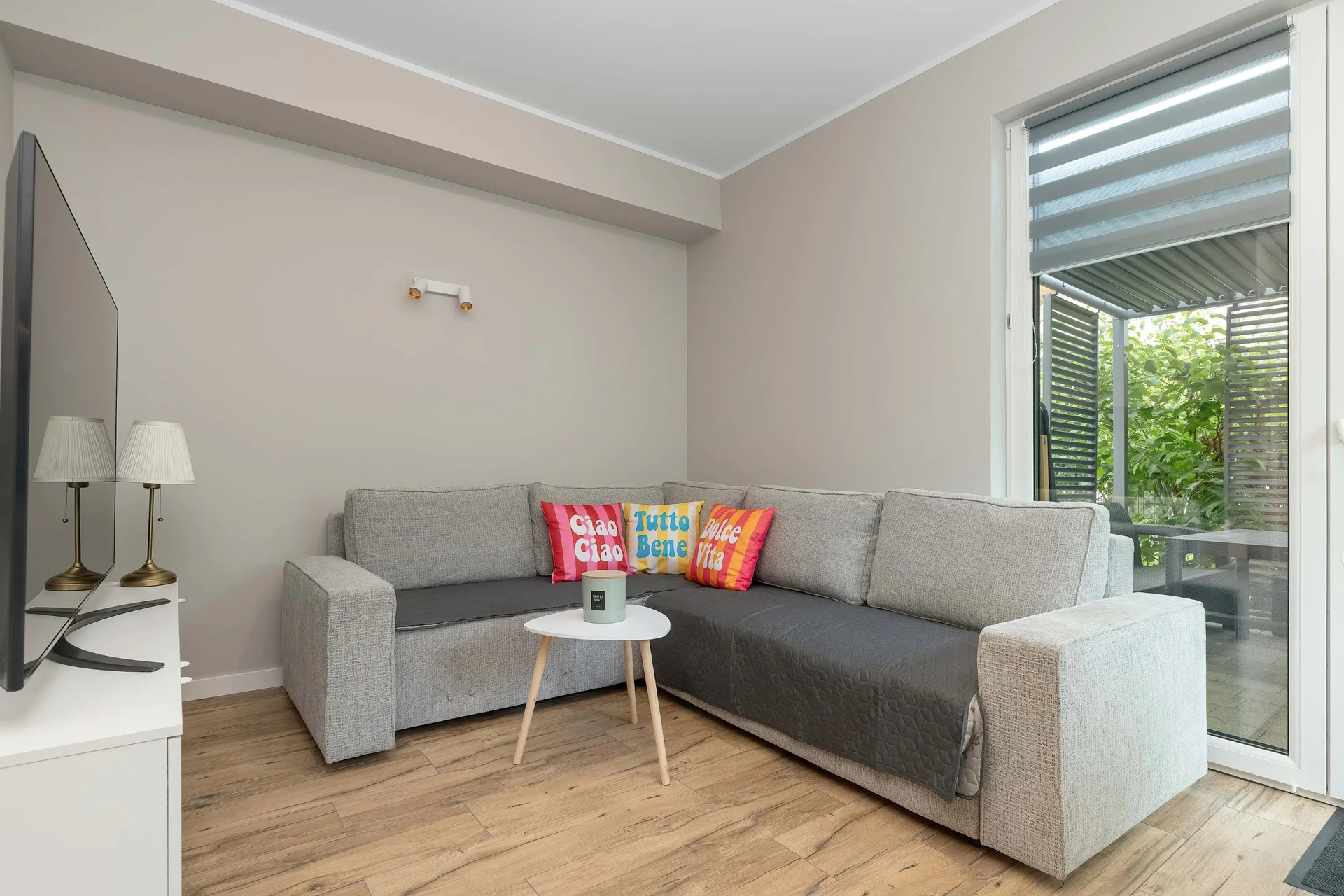 A bright living area highlights a spacious grey corner sofa placed on light laminate floors. Wide sliding glass doors open directly toward the verdant garden patio, ensuring smooth indoor-outdoor flow and abundant daylight.