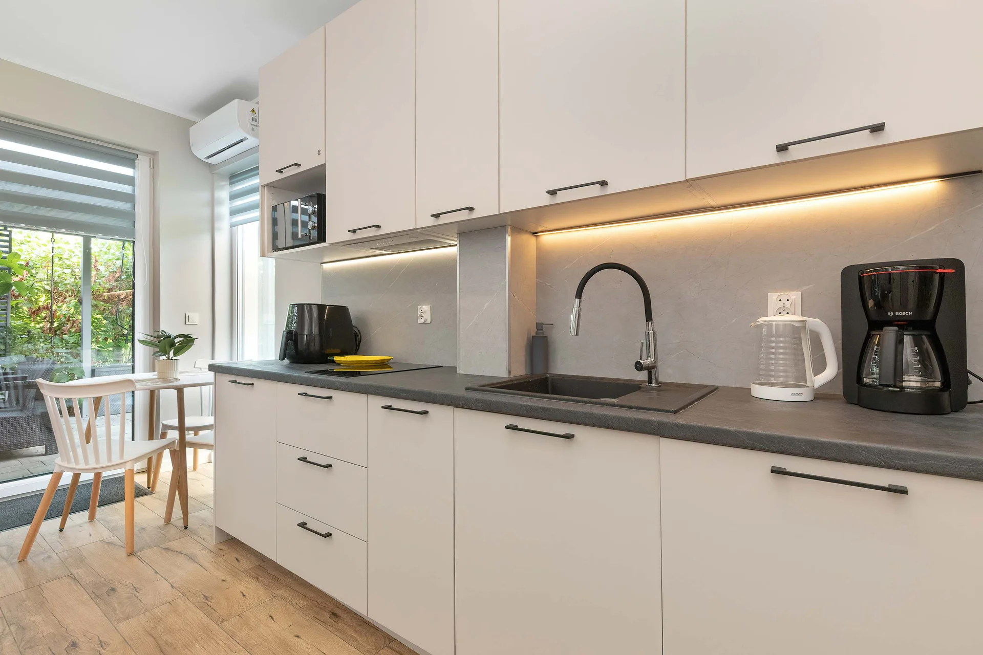 A contemporary open kitchenette offers sleek white handleless cabinets paired with dark countertops and under-cabinet lighting. Practical appliances include an electric kettle and coffee maker. Large windows brighten the room.