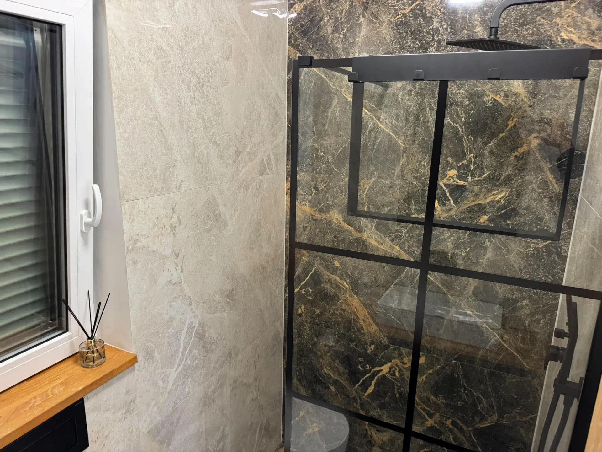 A modern bathroom interior highlights a walk-in shower enclosed with black-framed glass and marble-style tiles. A narrow window sits adjacent to light, textured walls. Clean fixtures create a contemporary and functional aesthetic.