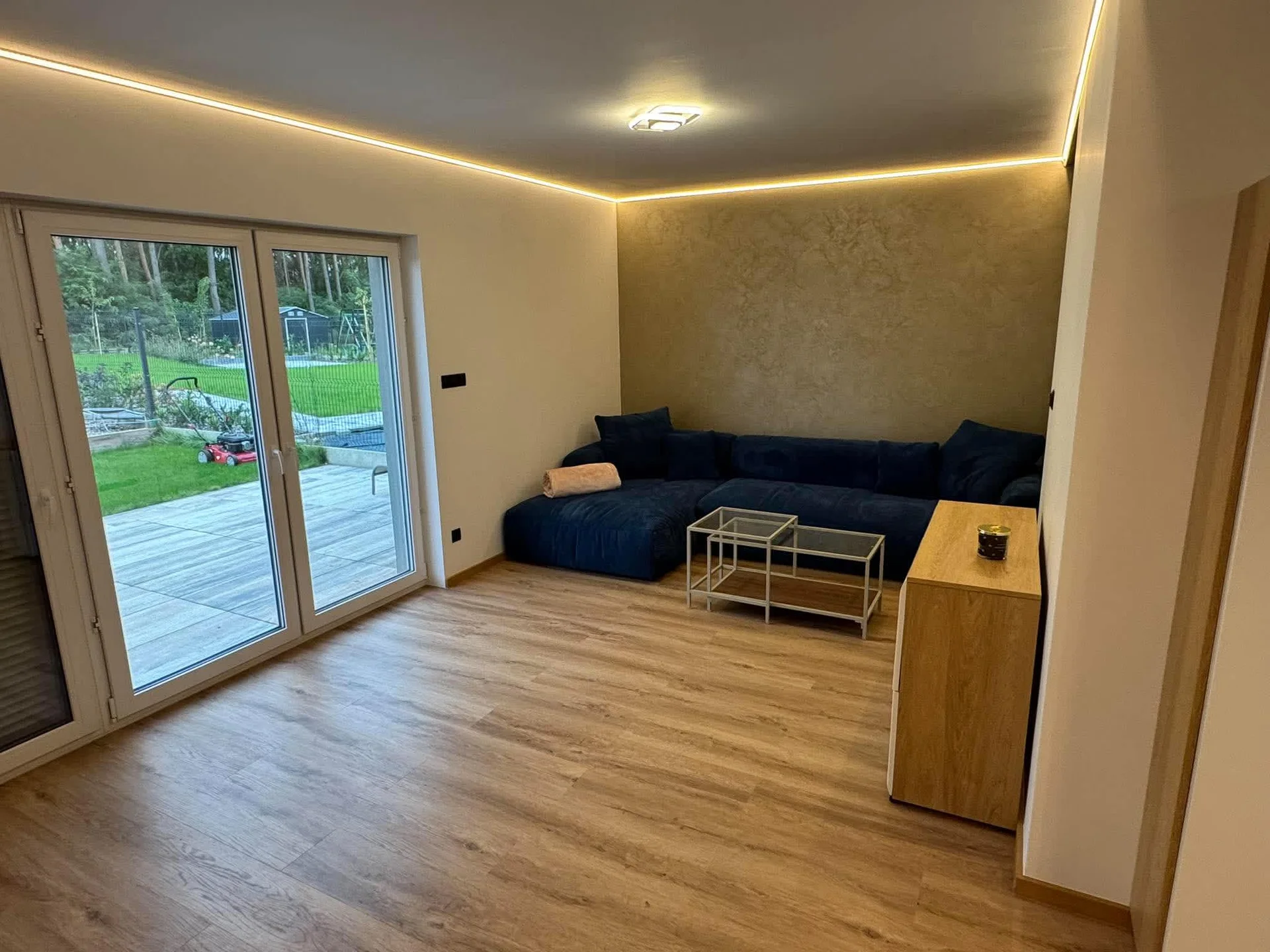 A bright living room features light wood floors and large sliding glass patio doors. A navy blue corner sofa rests beneath warm cove ceiling lighting. A coffee table and wooden credenza complete the open layout.