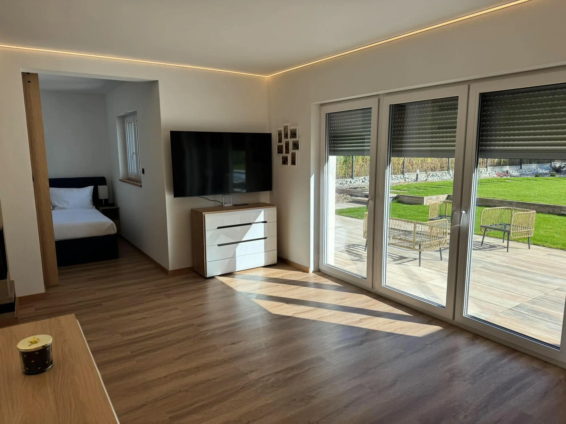 An open-plan living area displays wood laminate flooring and broad glass terrace doors. In the background, a doorway leads to a bedroom, while a TV hangs over a low cabinet. The spacious layout connects seamlessly to the outdoors.