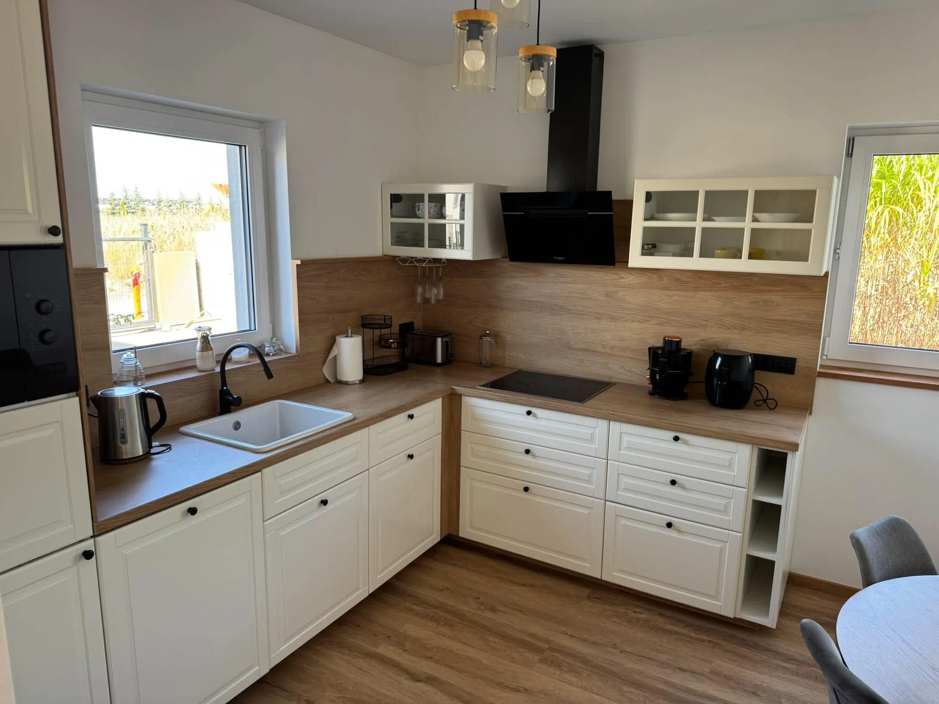 A modern L-shaped kitchen offers white base cabinetry paired with light wood countertops. A black range hood is mounted above the stovetop between upper cabinets. Sunlight streams in through a window directly over the sink.