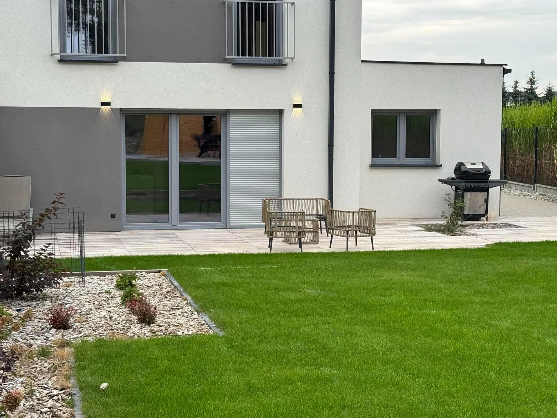 The exterior back garden features a green lawn bordered by decorative gravel and stepping stones. Patio furniture is arranged on the stone terrace next to the modern house. Glass doors provide direct ground-floor access outside.