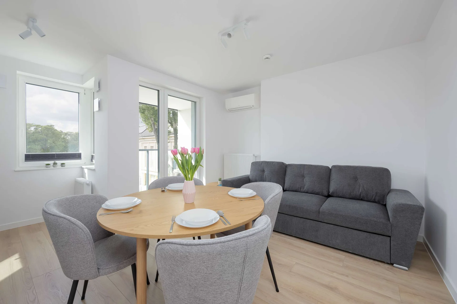 Bright living room with a grey sofa, round dining table, chairs and large windows providing plenty of natural light.
