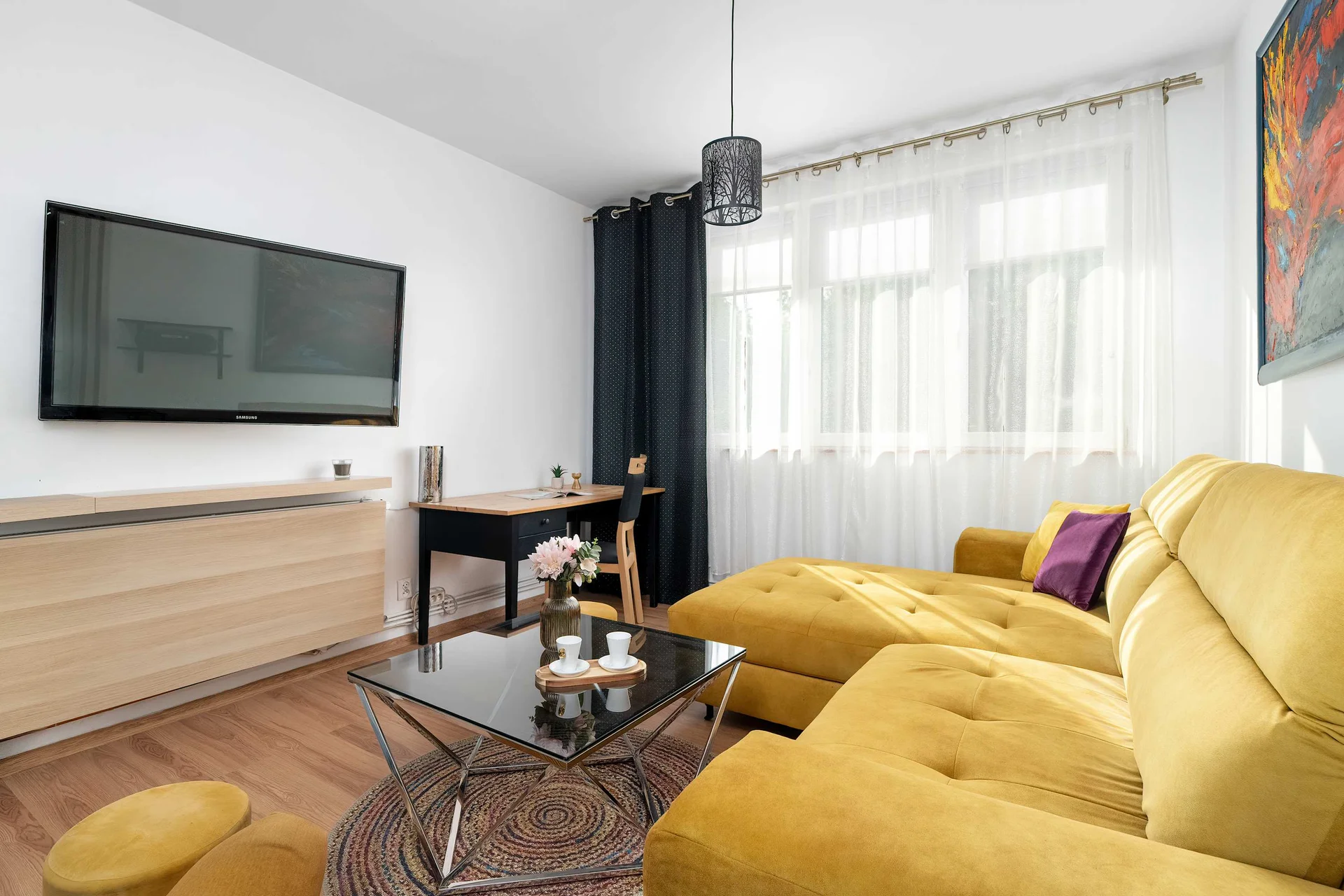 A bright living room with a mustard-yellow sofa, wall-mounted TV, wooden media cabinet, and a glass coffee table.
